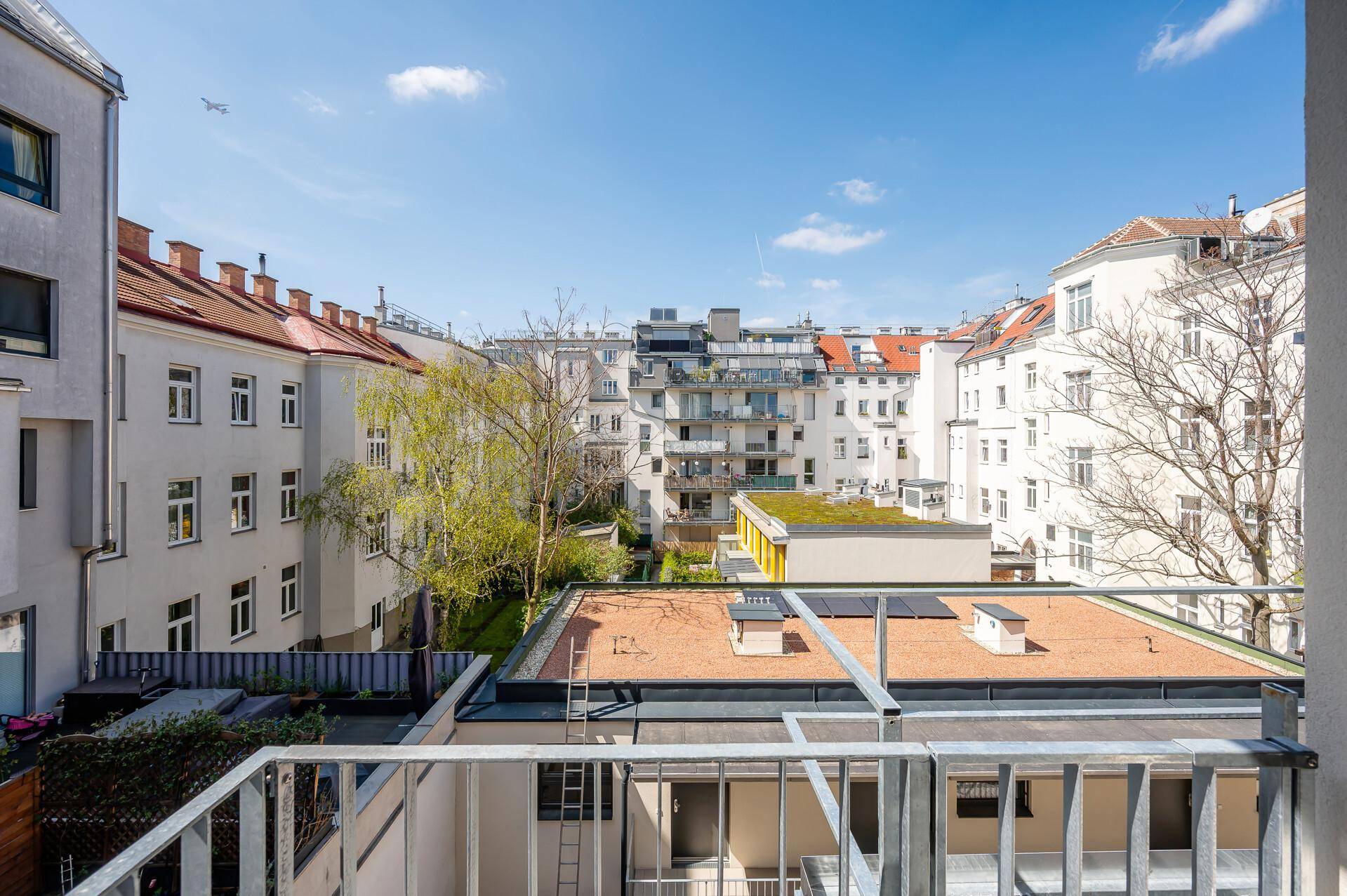 Blick vom Balkon in einen ruhigen Innenhof mit umliegenden Gebäuden, Bäumen und begrünten Dächern.