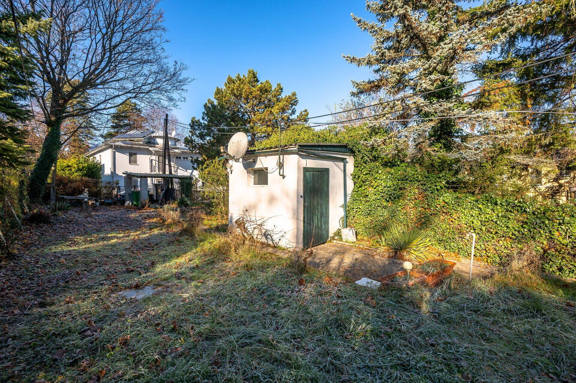 Kleiner Gartenschuppen mit Satellitenschüssel, umgeben von Bäumen und Sträuchern im Garten.