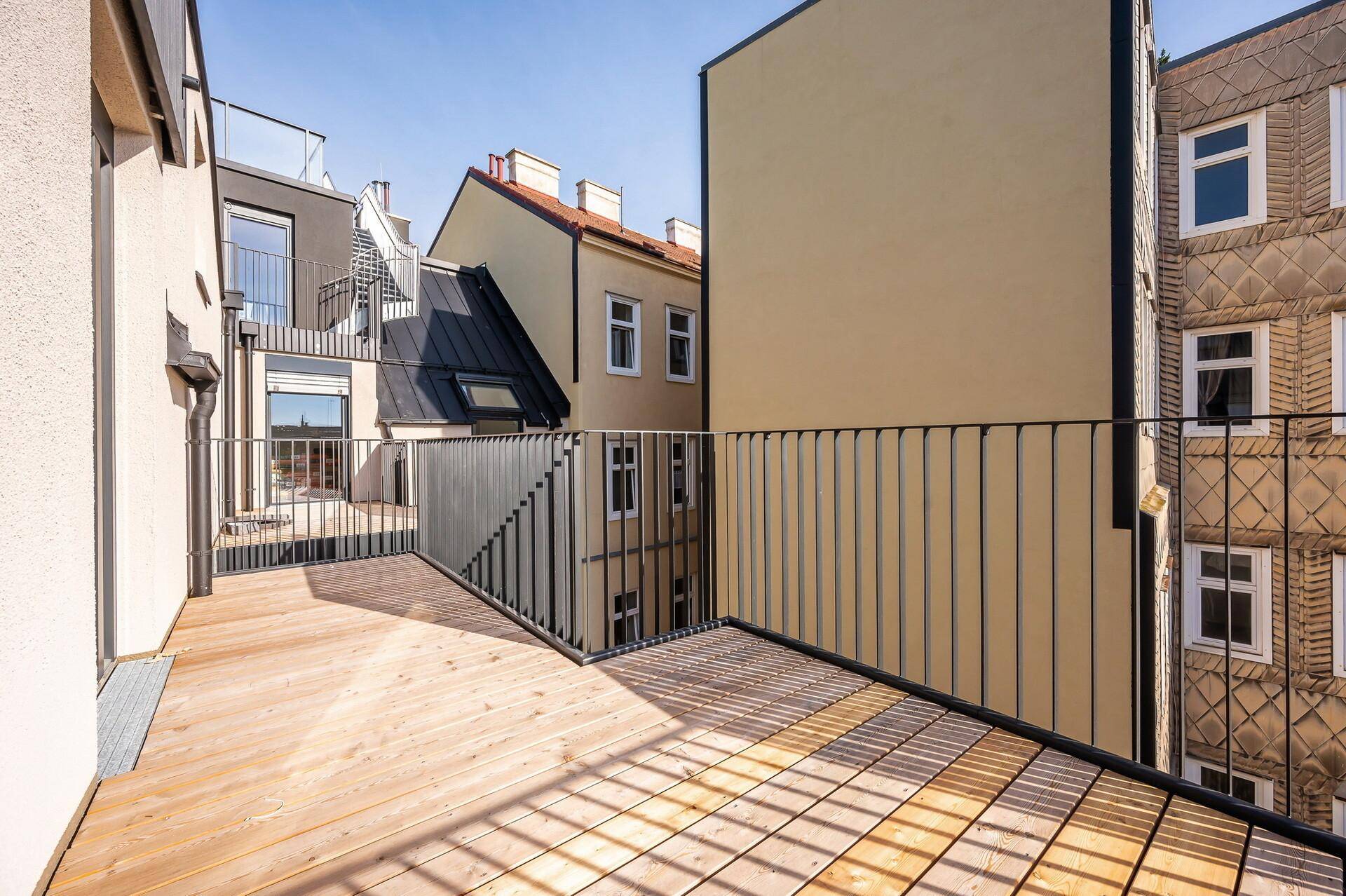 Geräumiger Balkon mit Holzboden und Blick auf die Nachbargebäude unter blauem Himmel.