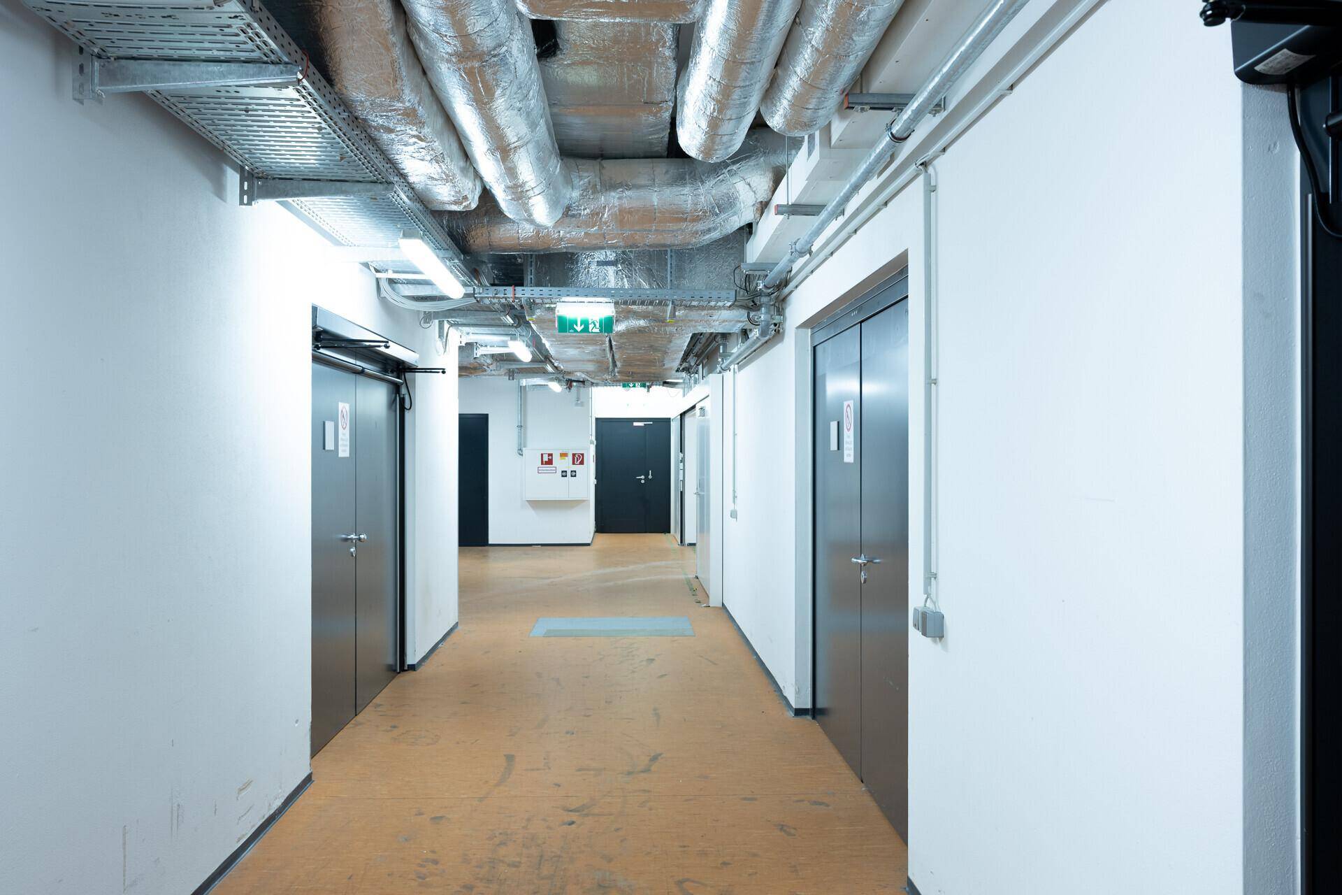 Industrieller Flur mit freiliegenden Rohren und Lüftungskanälen an der Decke sowie mehreren Türen und Aufzügen.
