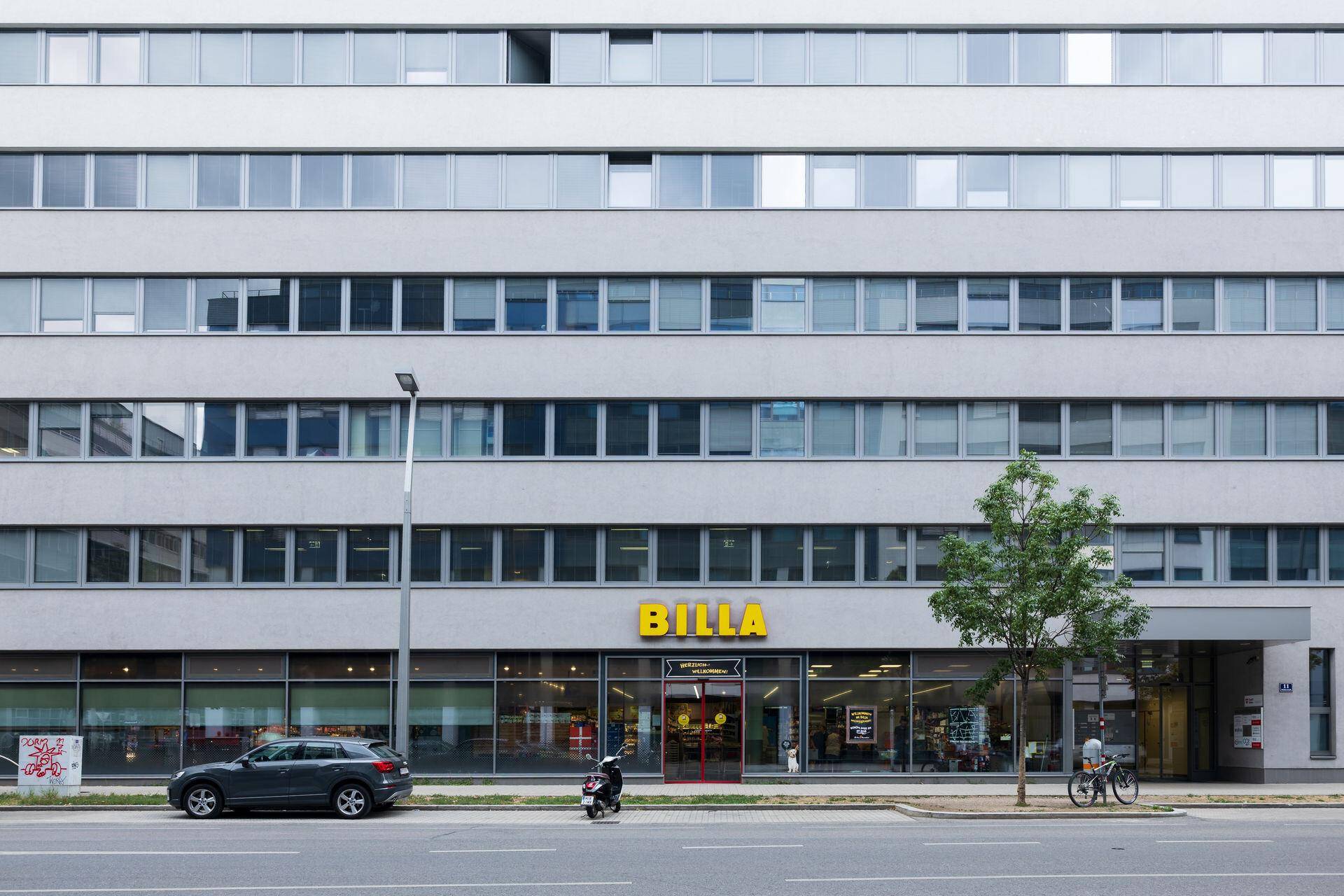 Straßenseite des Gebäudes mit einem BILLA Supermarkt im Erdgeschoss und parkenden Autos.