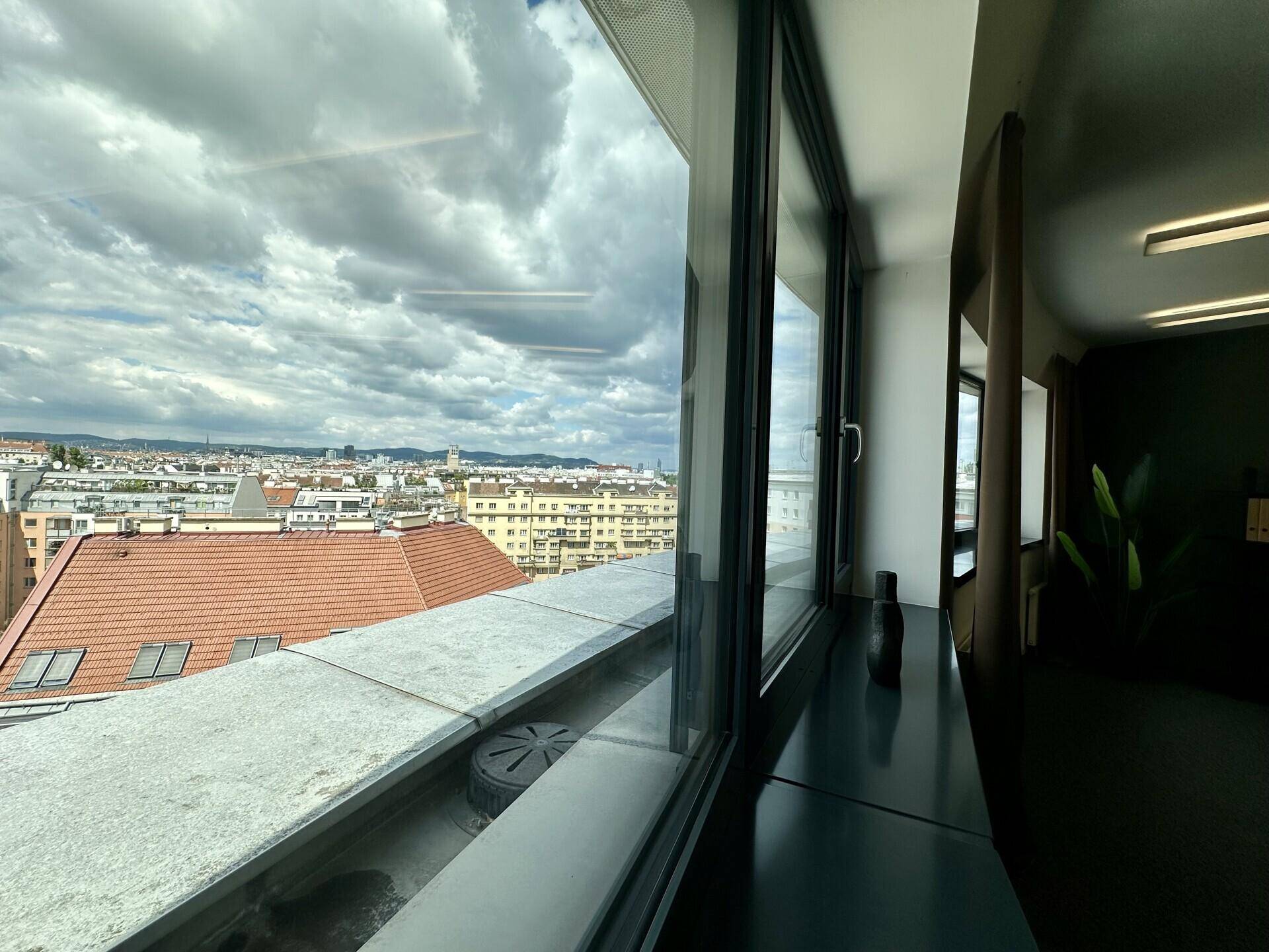 Innenraum mit großen Fenstern, die einen weiten Blick über die Stadt und den Himmel bieten.
