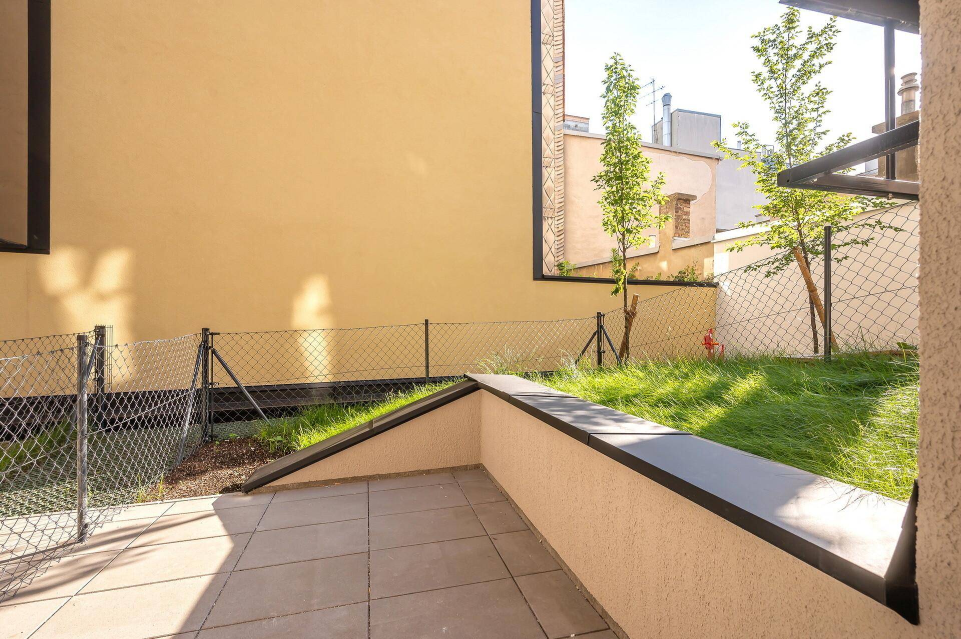 Sonniger Außenbereich mit gepflasterter Terrasse und kleiner Rasenfläche, umgeben von einem Zaun.
