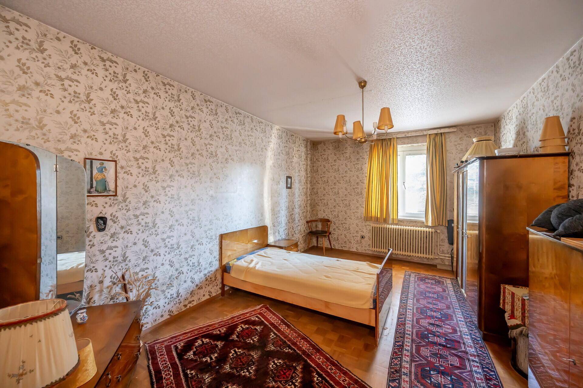 Schlafzimmer mit floraler Tapete, Holzmöbeln und einem Fenster mit Blick ins Grüne.