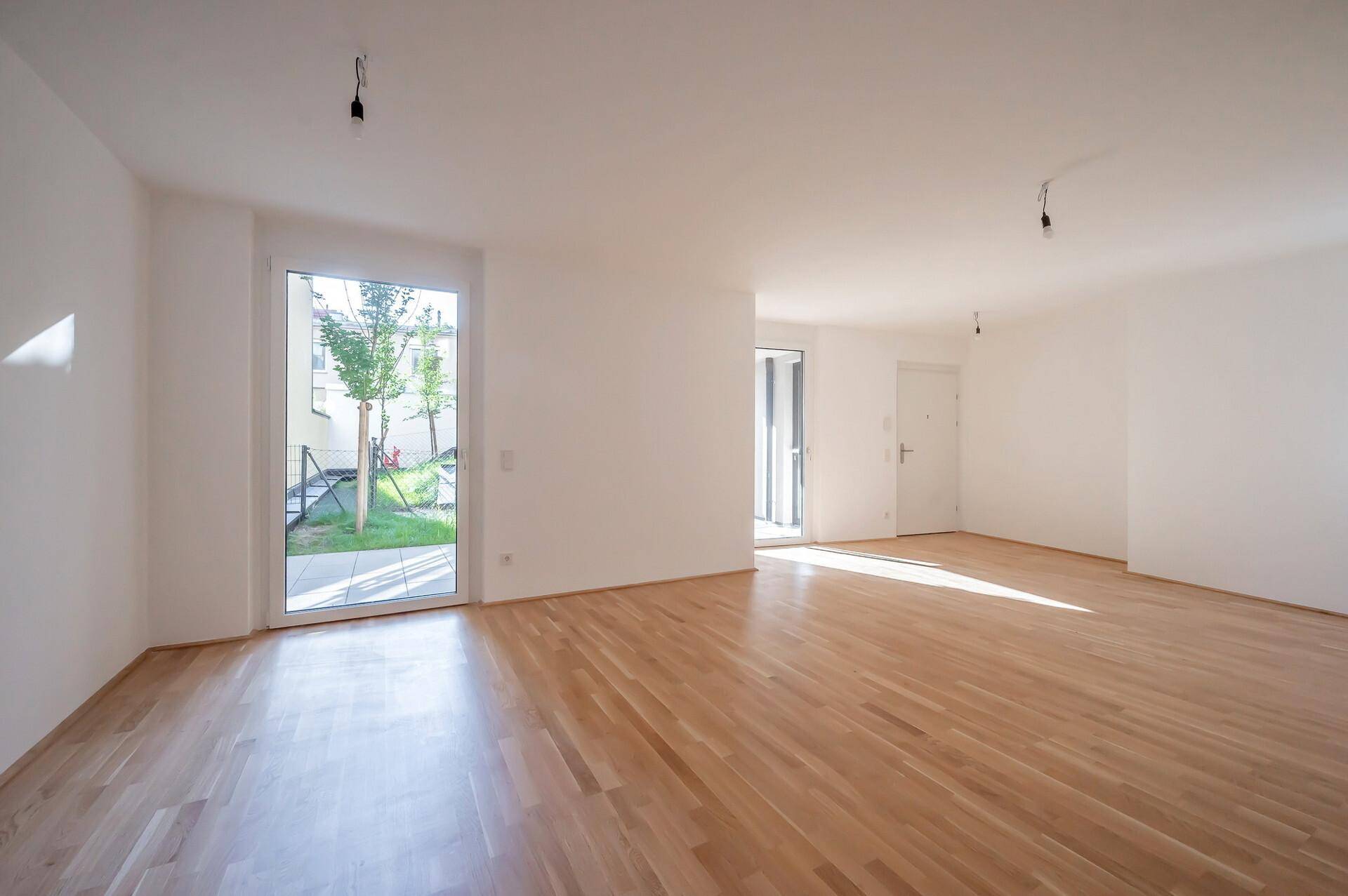 Ein heller, leerer Wohnraum mit Holzboden und großen Glastüren, die viel Tageslicht hereinlassen.