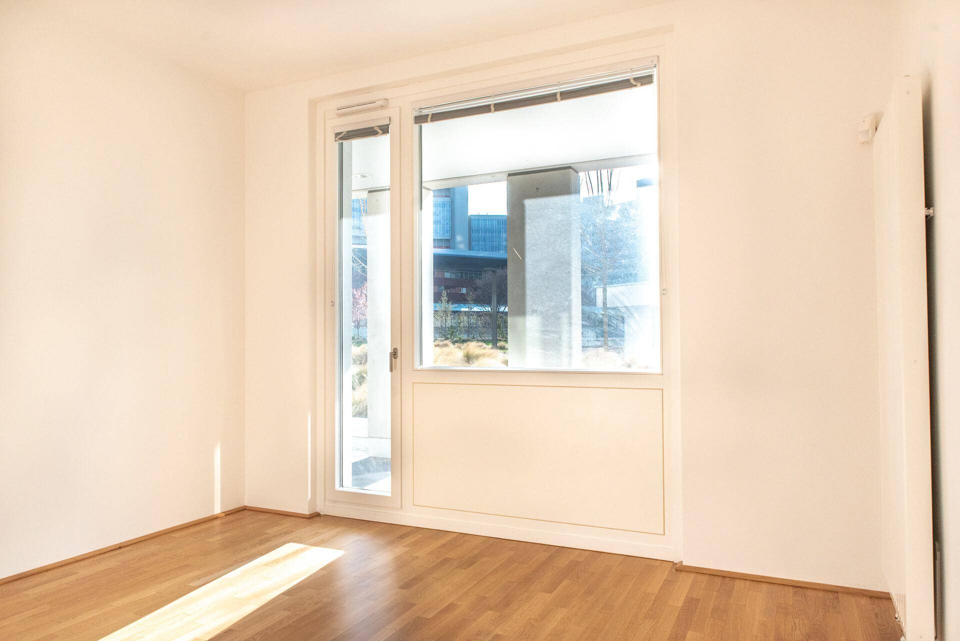 Helles Schlafzimmer mit Holzboden und großem Fenster, das viel Sonnenlicht hereinlässt.