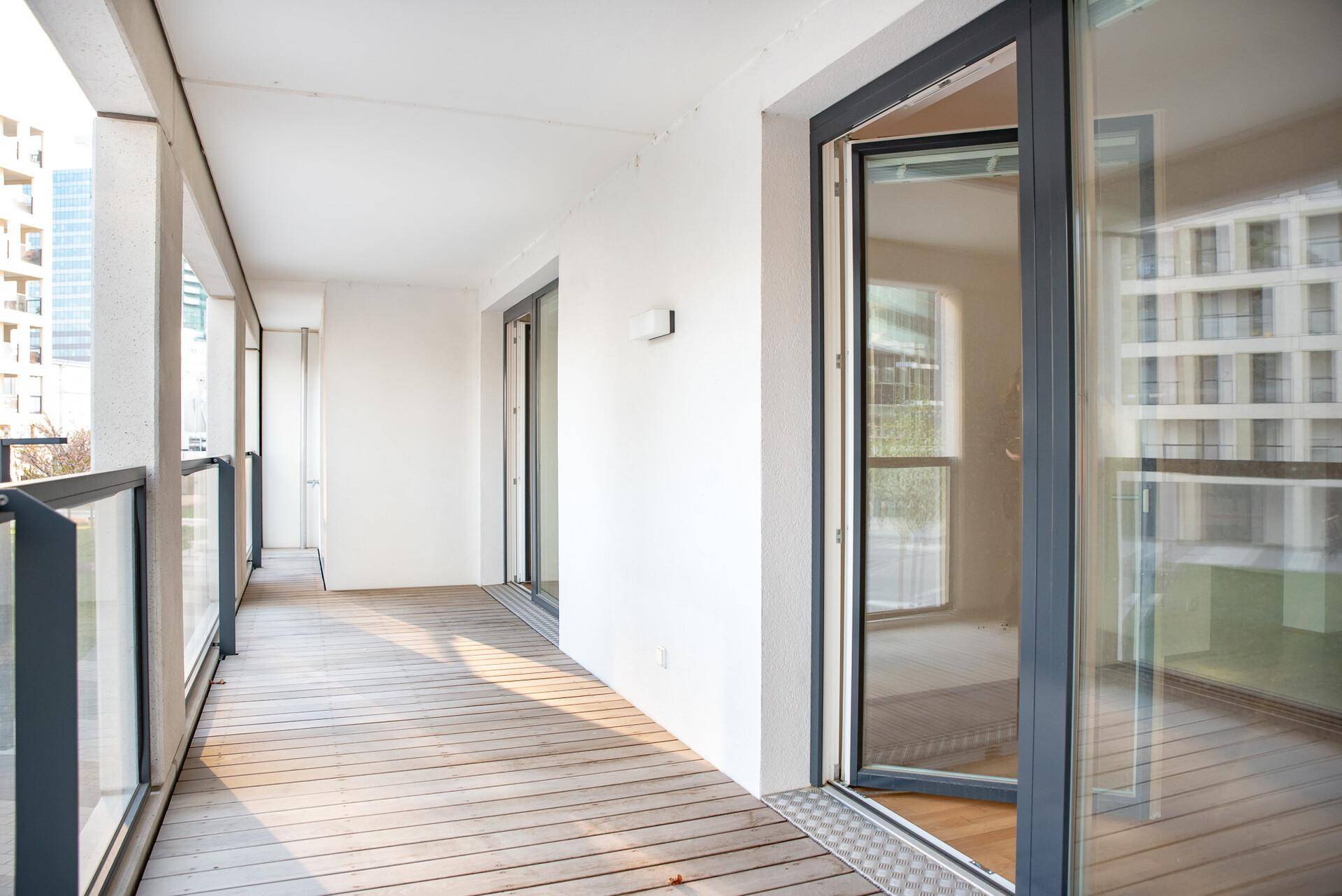 Langer Balkon mit Holzdielenboden und Glasgeländer, bietet Blick auf die umliegenden Gebäude.