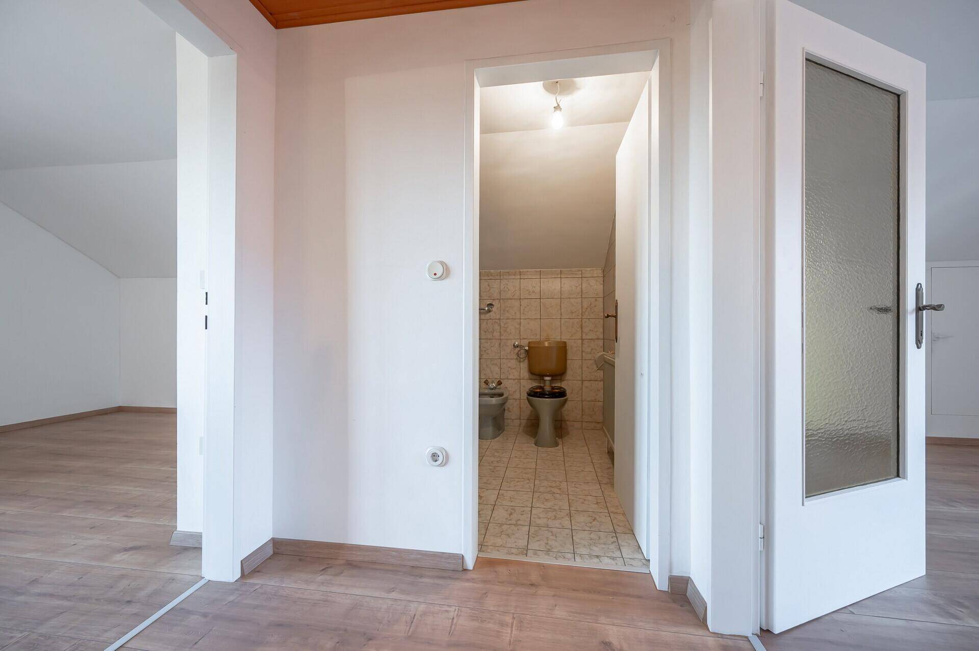Heller Flur mit Laminatboden, der zu einem Badezimmer mit älterer Ausstattung führt.
