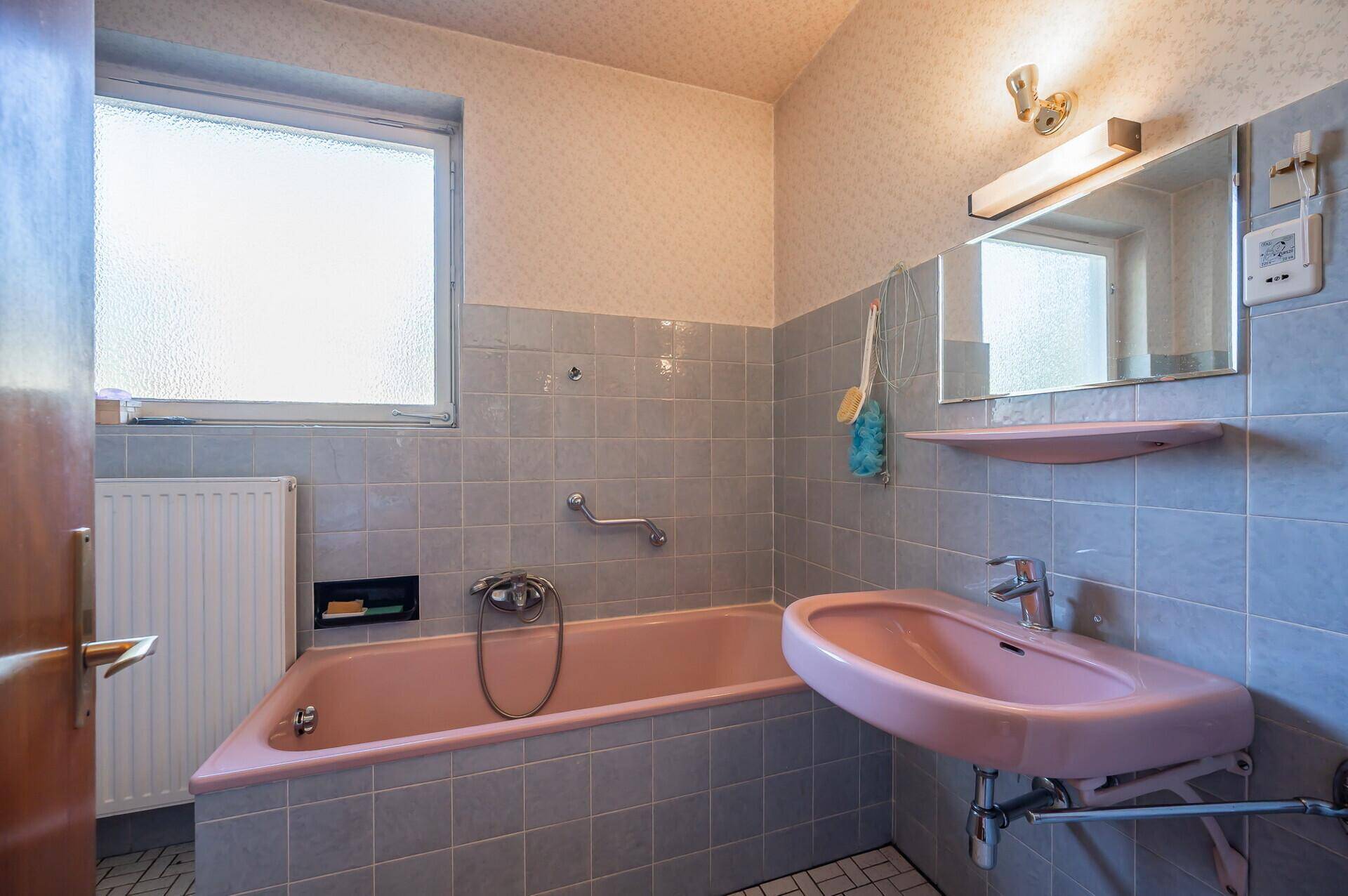 Badezimmer im 80er-Jahre-Stil mit rosa Badewanne, Waschbecken und grauen Fliesen.