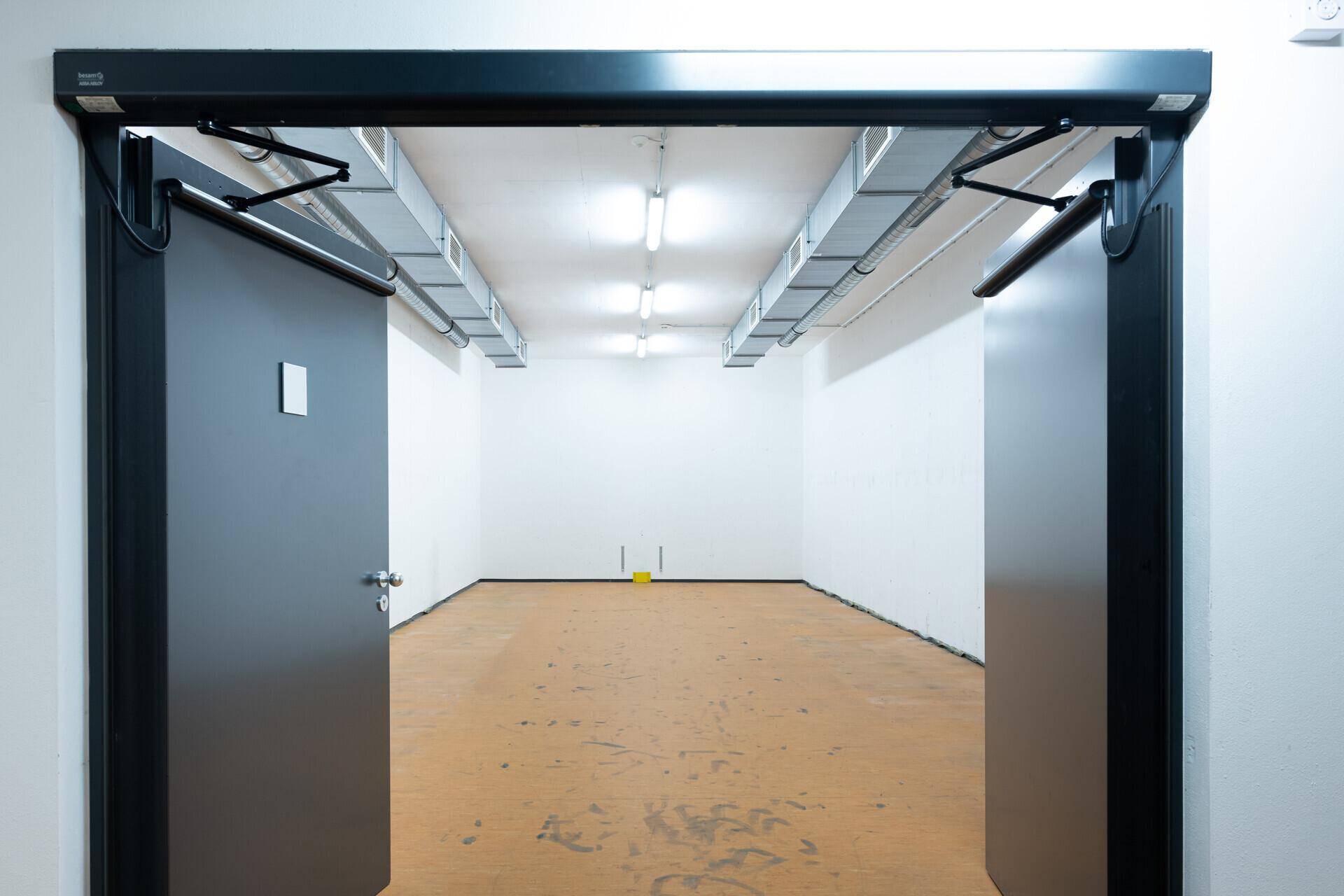 Leerer Raum mit weißen Wänden, braunem Boden und sichtbaren Lüftungskanälen an der Decke, gesehen durch eine offene Tür.