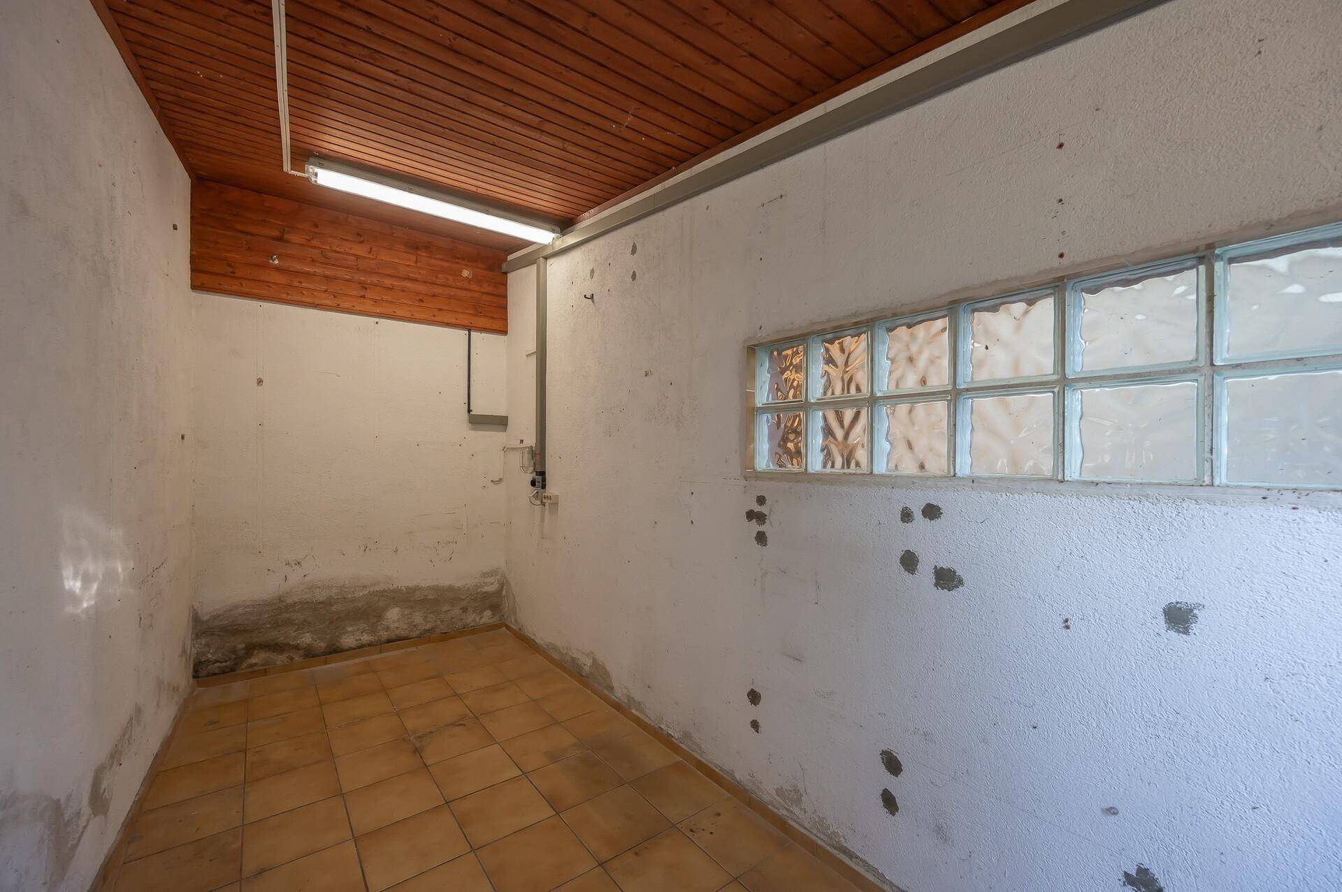 Funktionaler Abstellraum mit Fliesenboden, Holzdecke und Fenster aus Glasbausteinen.
