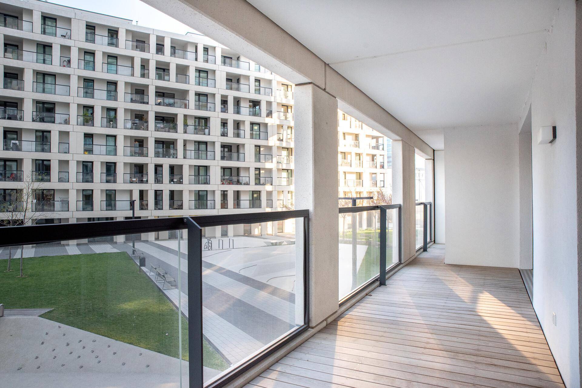 Breiter Balkon mit Holzdielen und Glasgeländer, mit Blick auf einen begrünten Innenhof.