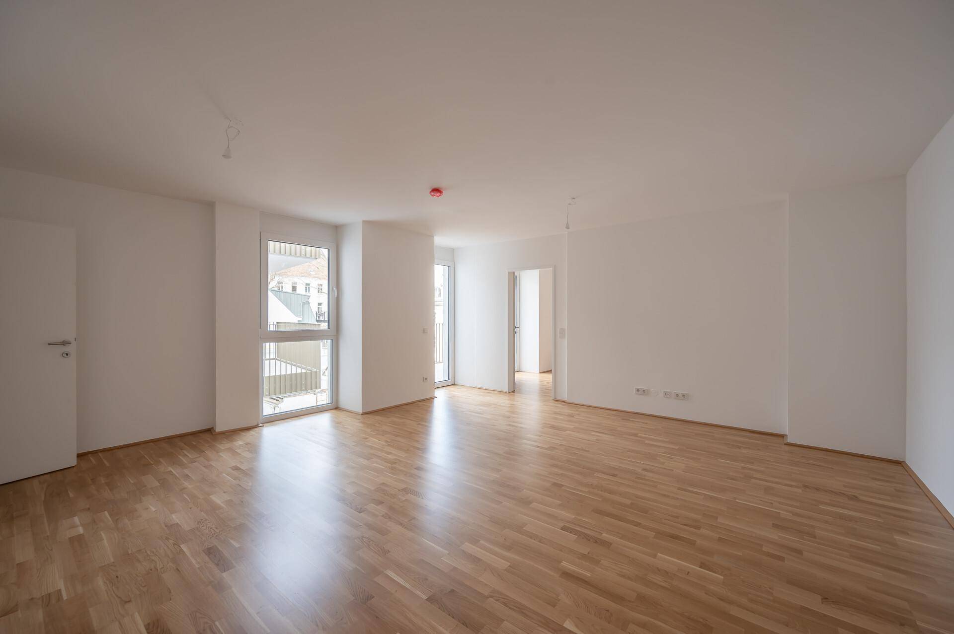 Heller, leerer Wohnbereich mit Parkettboden und großen Fenstern, die viel Tageslicht hereinlassen.