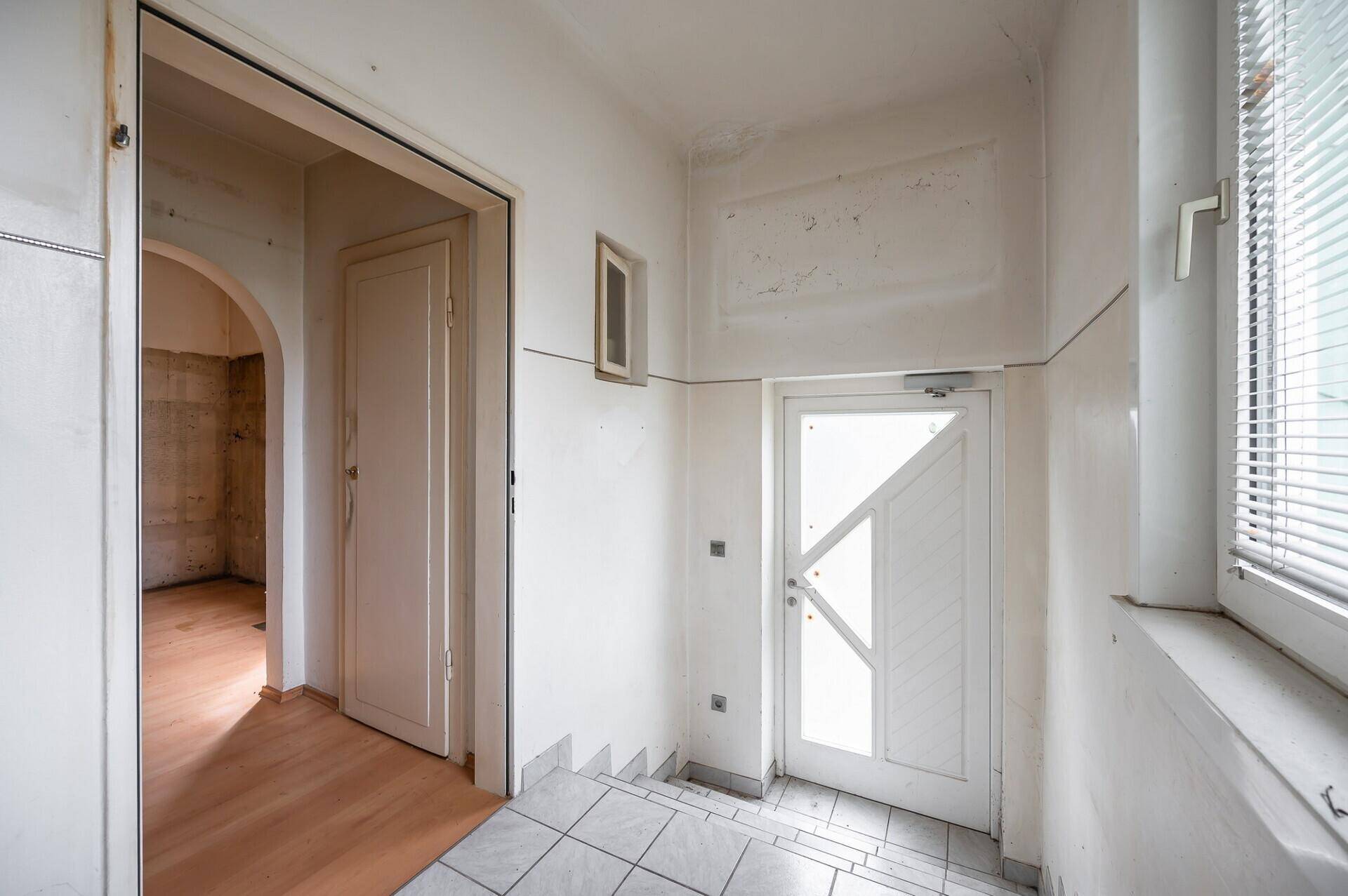 Eingangsbereich mit Fliesenboden, weißer Tür und Fenster mit Jalousien.