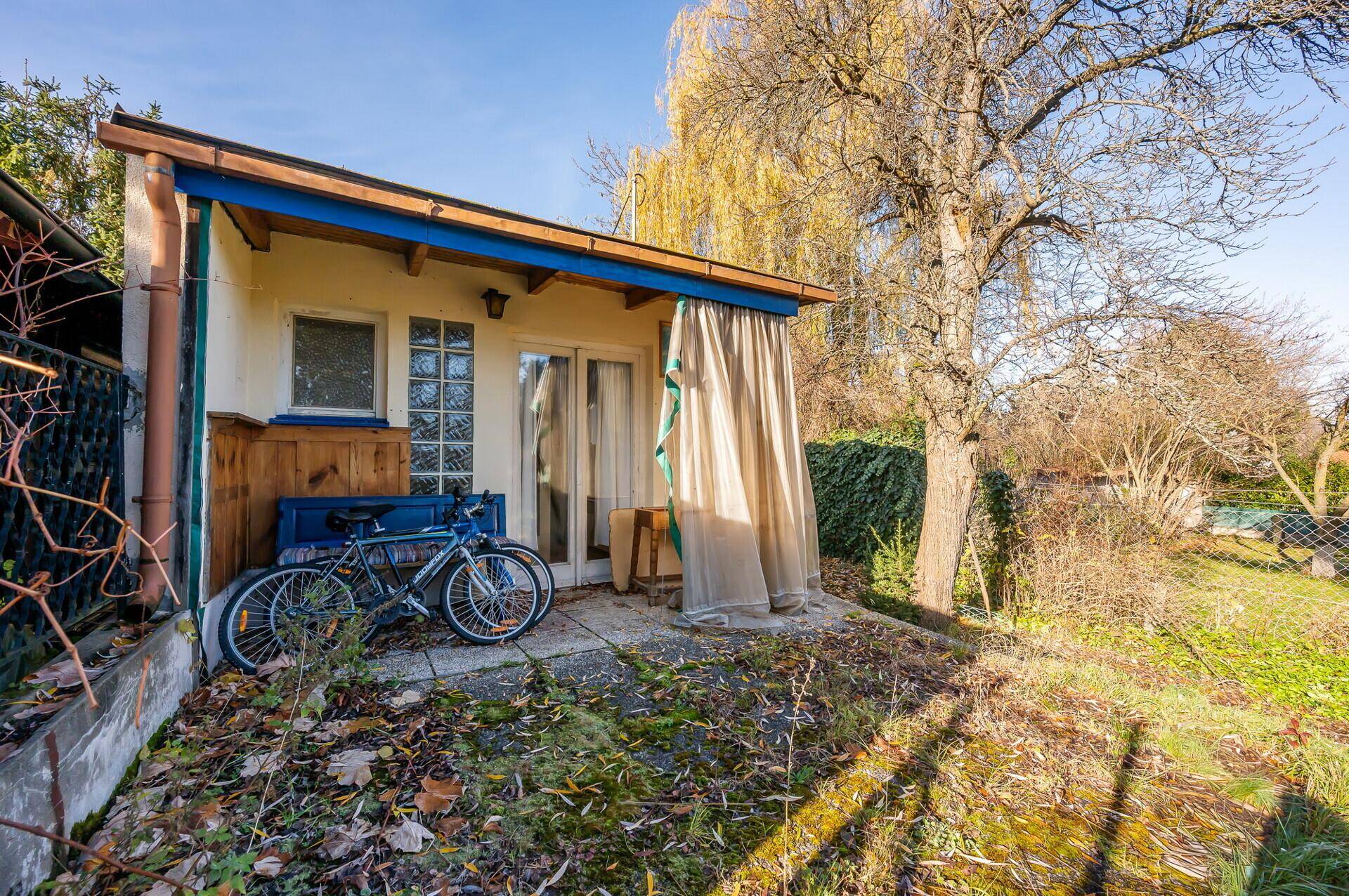 Kleine Terrasse am Gebäude mit Fahrrad und Vorhang, umgeben von herbstlicher Vegetation und Sonnenlicht.