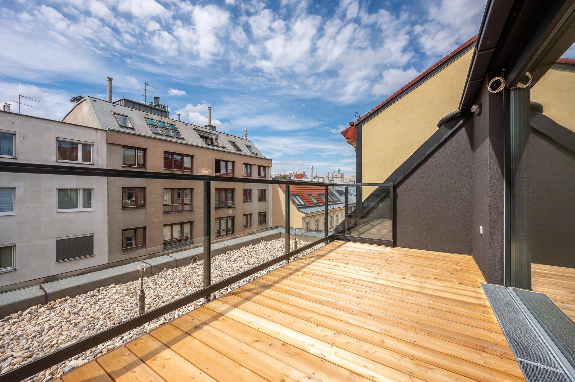 Sonniger Balkon mit Holzbelag und Glasgeländer bietet einen Blick auf die umliegenden Gebäude.