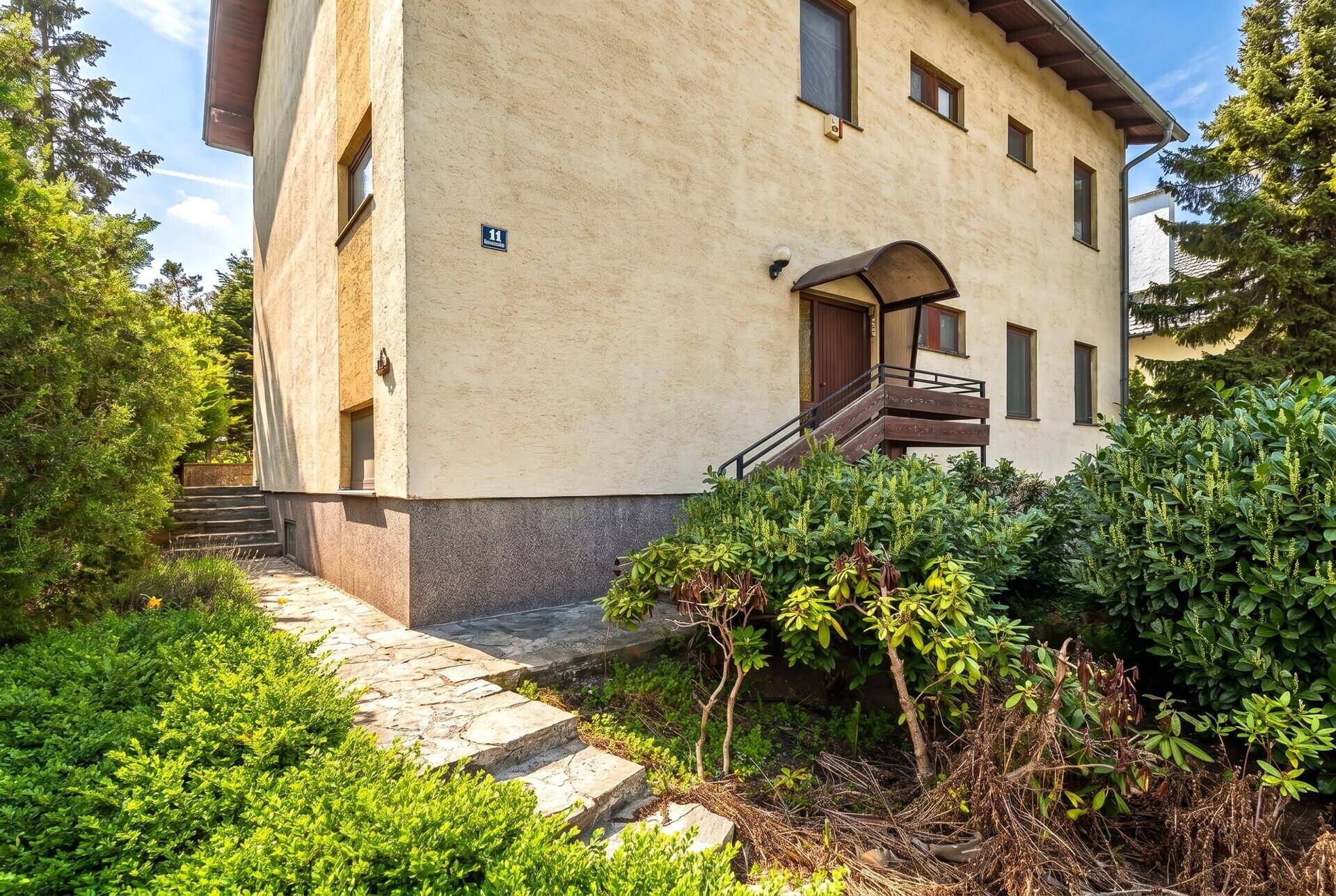 Seitenansicht des Hauses mit Eingangsbereich, Treppe und üppiger Bepflanzung im Vorgarten.