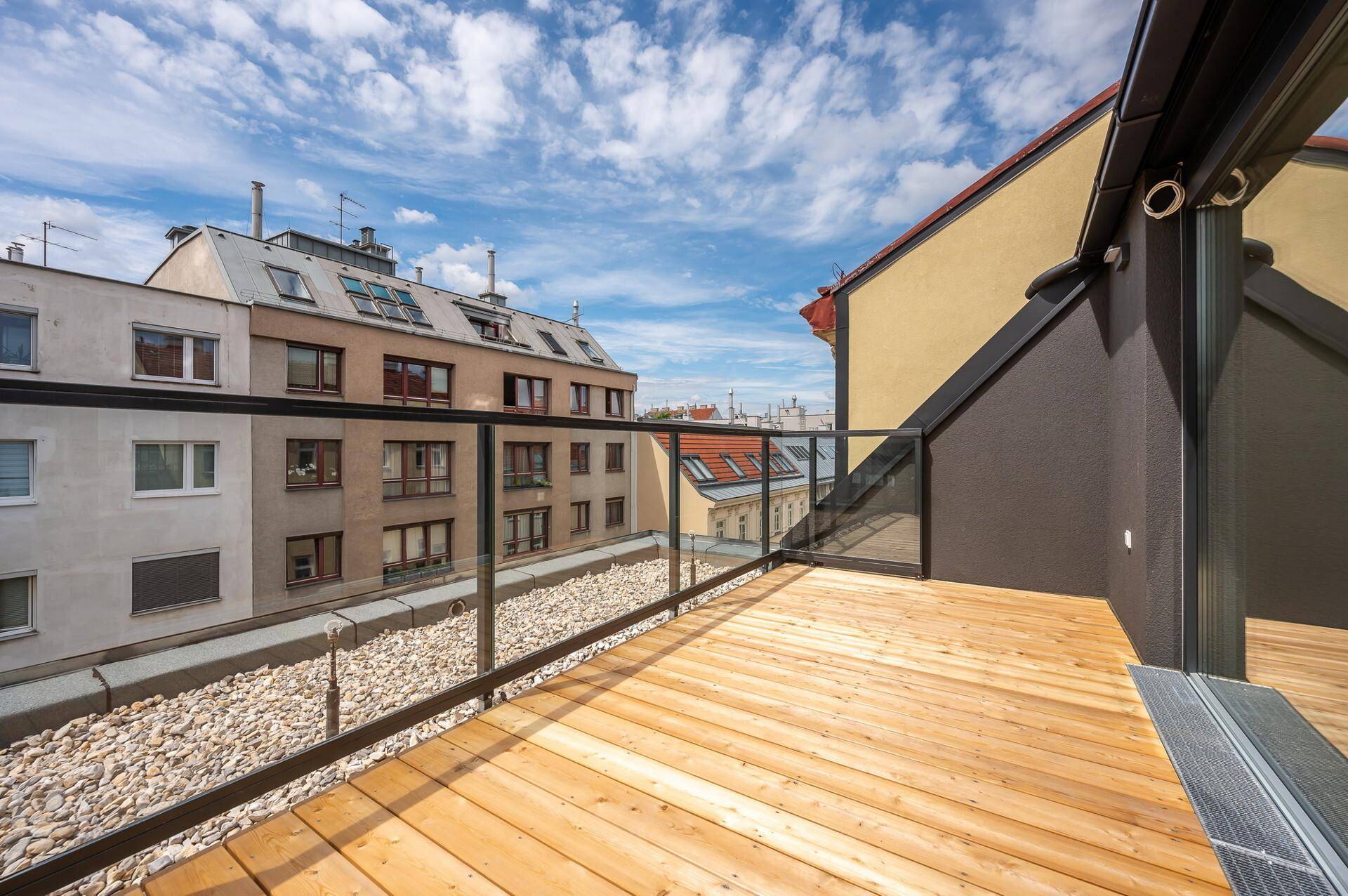Balkon mit Holzboden und Glasgeländer, bietet einen Blick auf die umliegenden Gebäude.