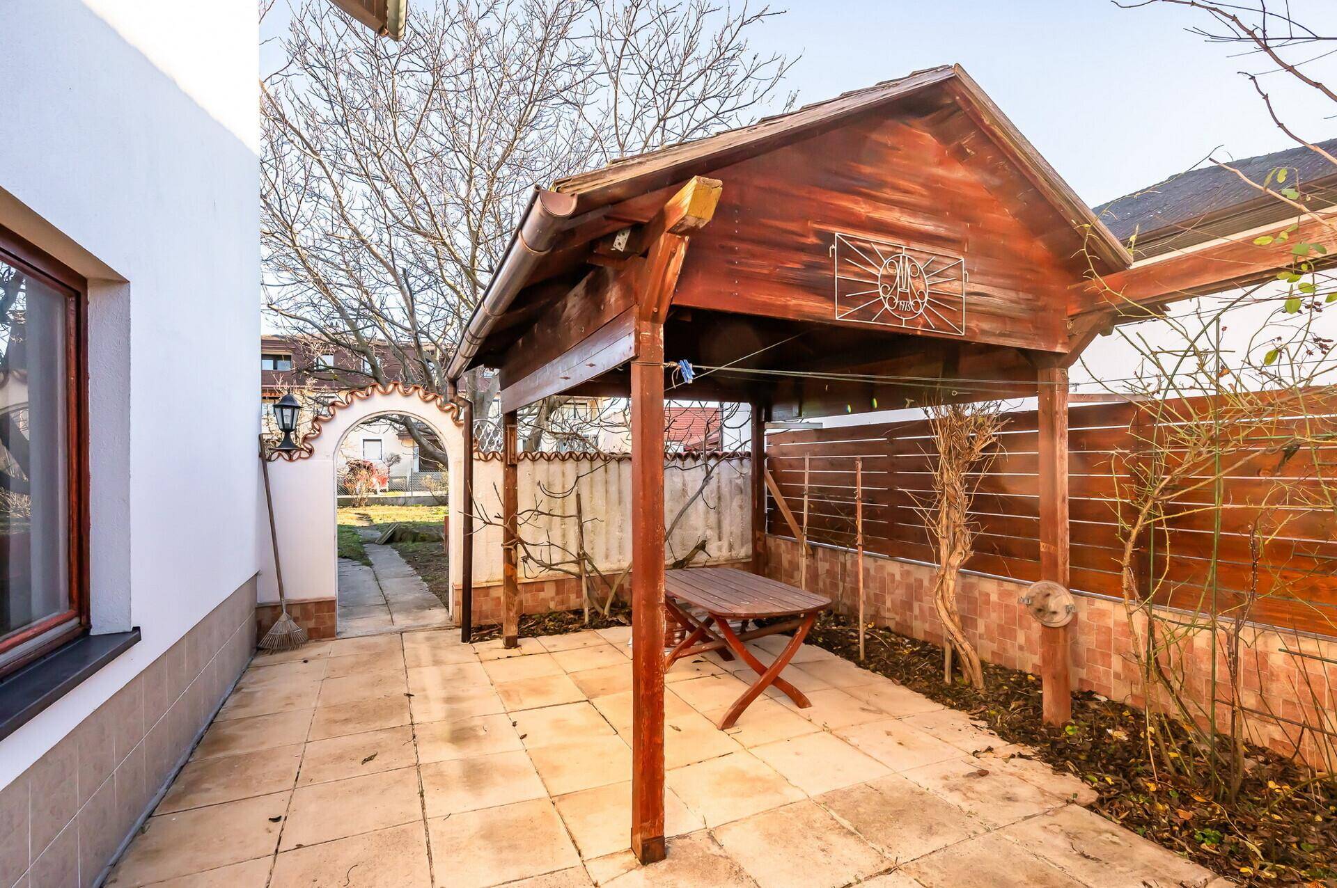 Überdachte Terrasse mit Holztisch und Pergola, umgeben von einem Holzzaun im Garten.