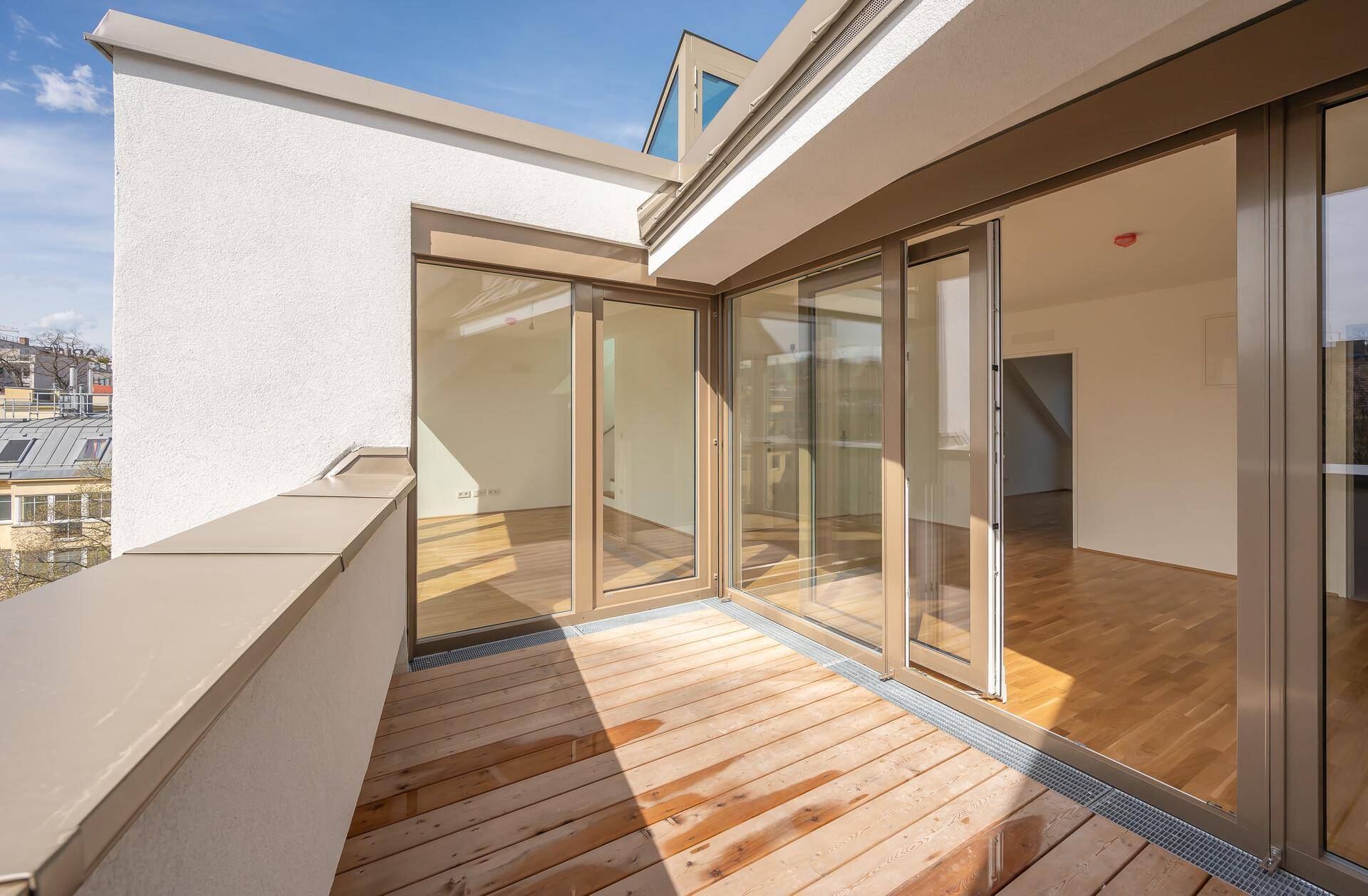 Moderner Balkon mit Holzdeck und großen Glasfronten, die den Innenraum mit Licht durchfluten.
