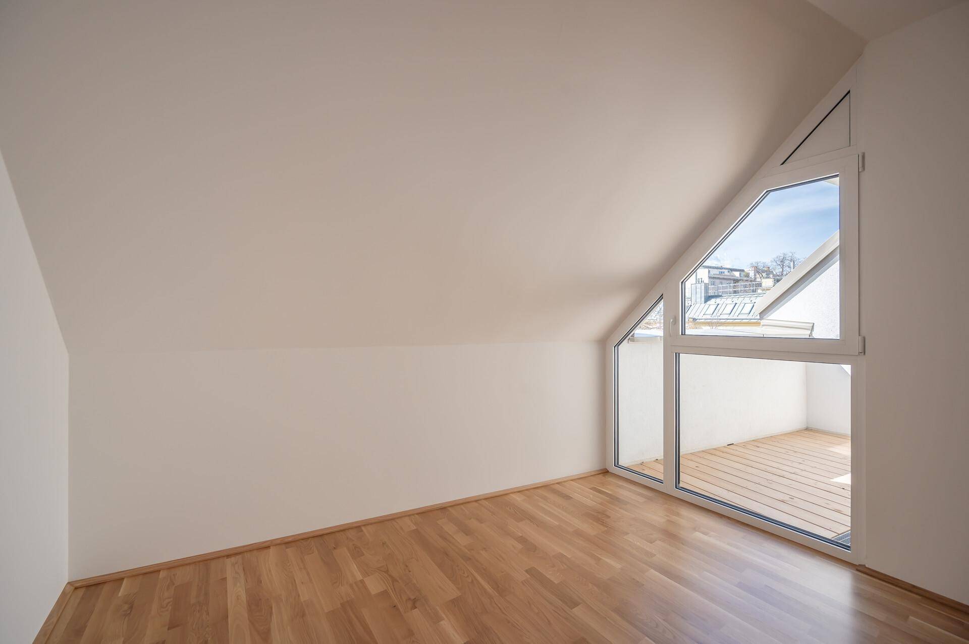 Heller Raum im Dachgeschoss mit schrägen Wänden, Holzboden und großen, schrägen Fenstern.