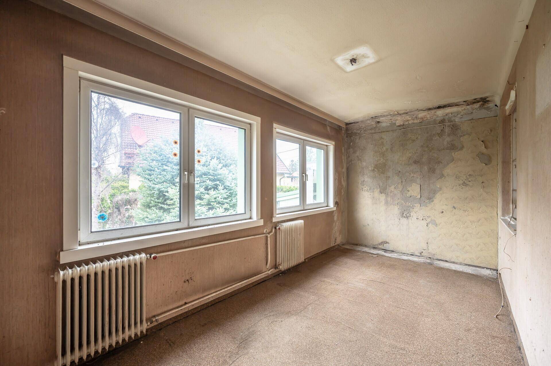 Geräumiger Raum mit Teppichboden, großen Fenstern und Heizkörper, renovierungsbedürftig.