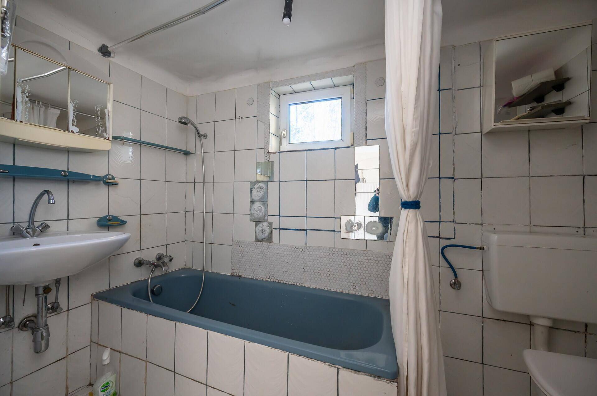 Badezimmer mit blauer Badewanne, Waschbecken und weißen Fliesen, renovierungsbedürftig.
