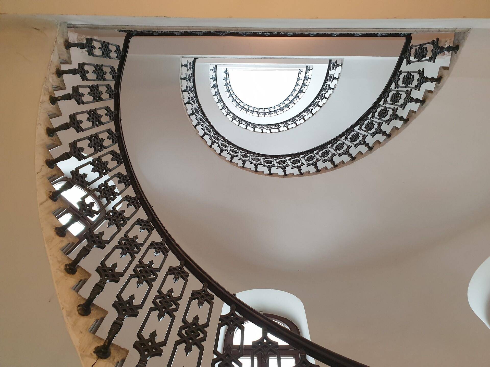 Blick nach oben in ein beeindruckendes, spiralförmiges Treppenhaus mit kunstvollem Geländer und Oberlicht.
