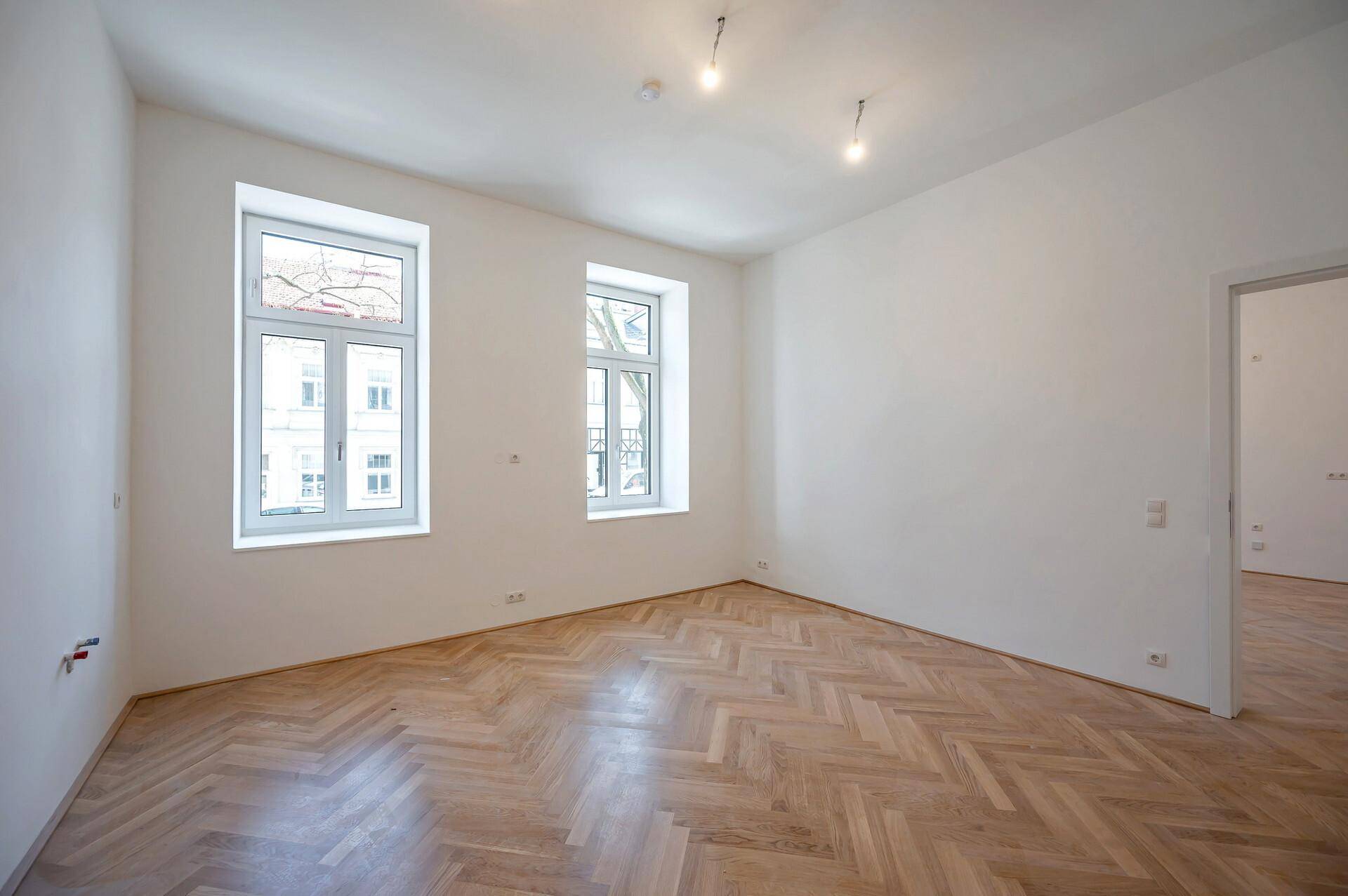 Geräumiger, unmöblierter Raum mit hellem Fischgrätparkett und Blick auf die Stadt durch die Fenster.