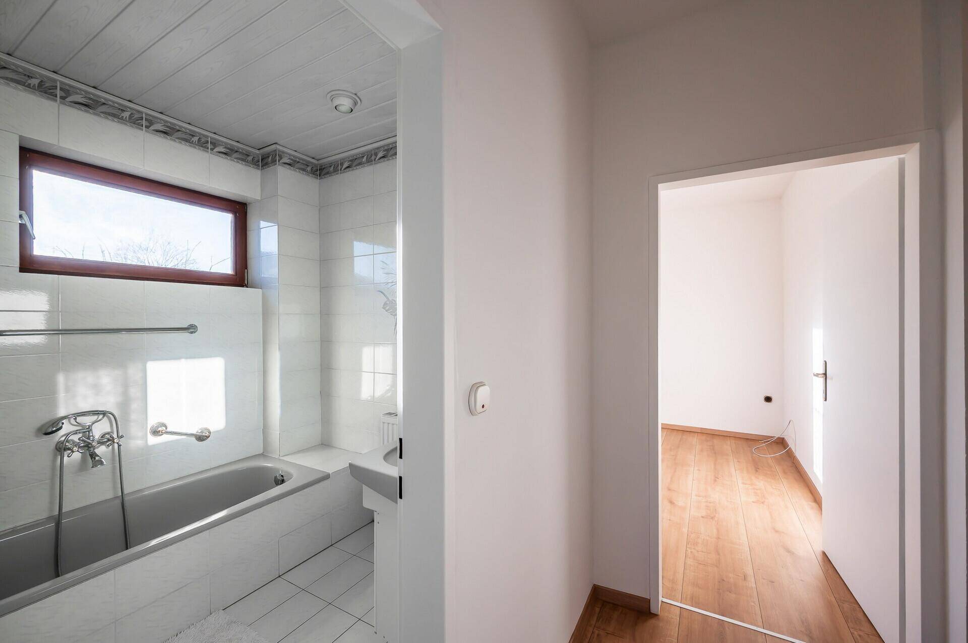 Badezimmer mit Badewanne, hellen Fliesen und einem Fenster, das Tageslicht hereinlässt.