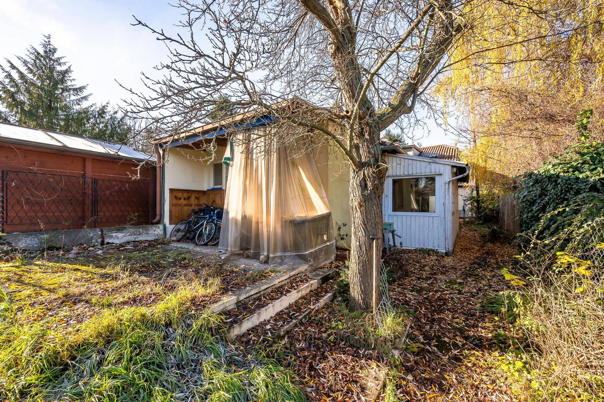 Kleines Gebäude im Garten mit Terrasse und umgebender Vegetation, renovierungsbedürftig.