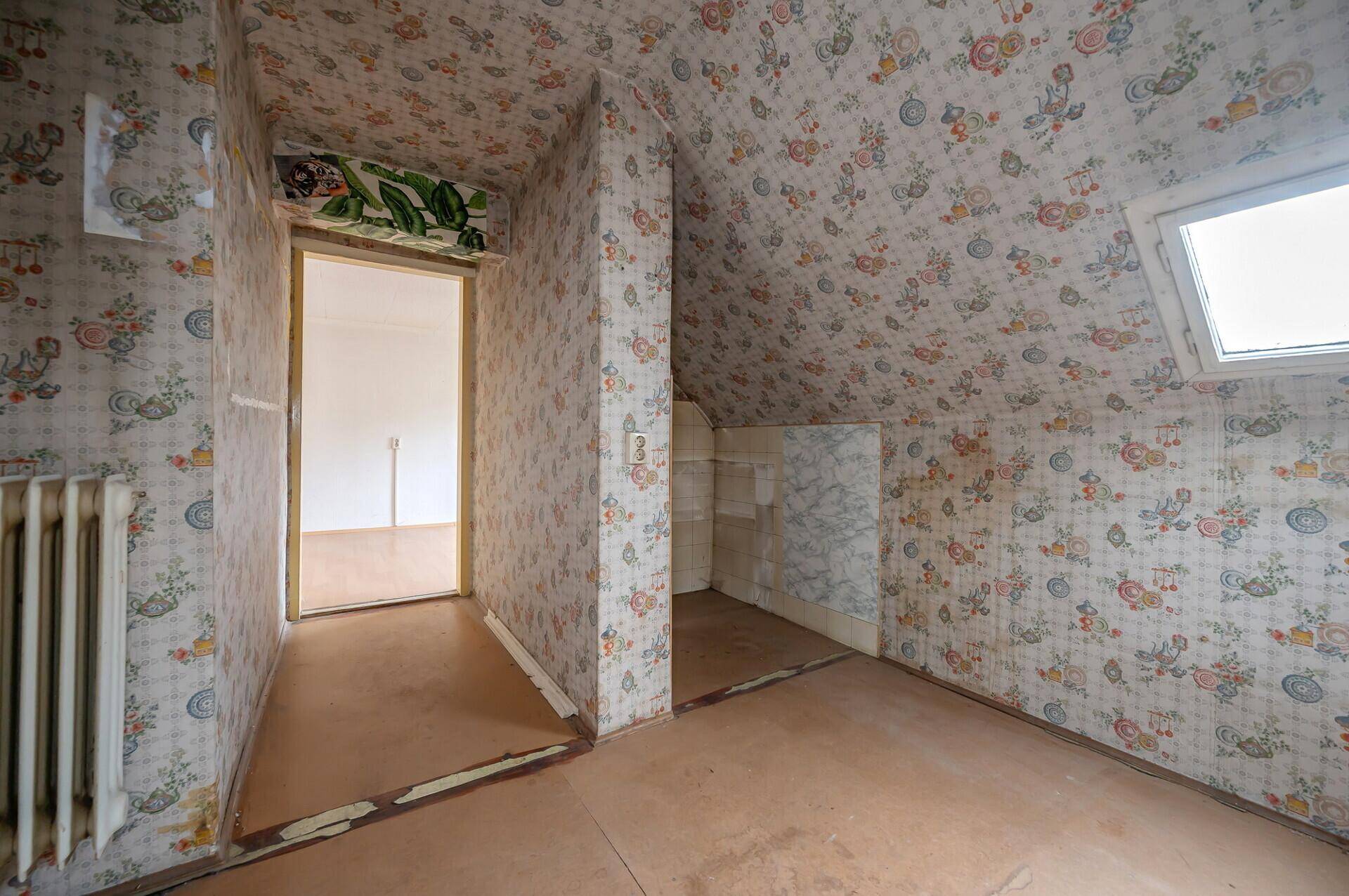 Dachgeschossraum mit schrägen Wänden, floraler Tapete und Dachfenster, renovierungsbedürftig.