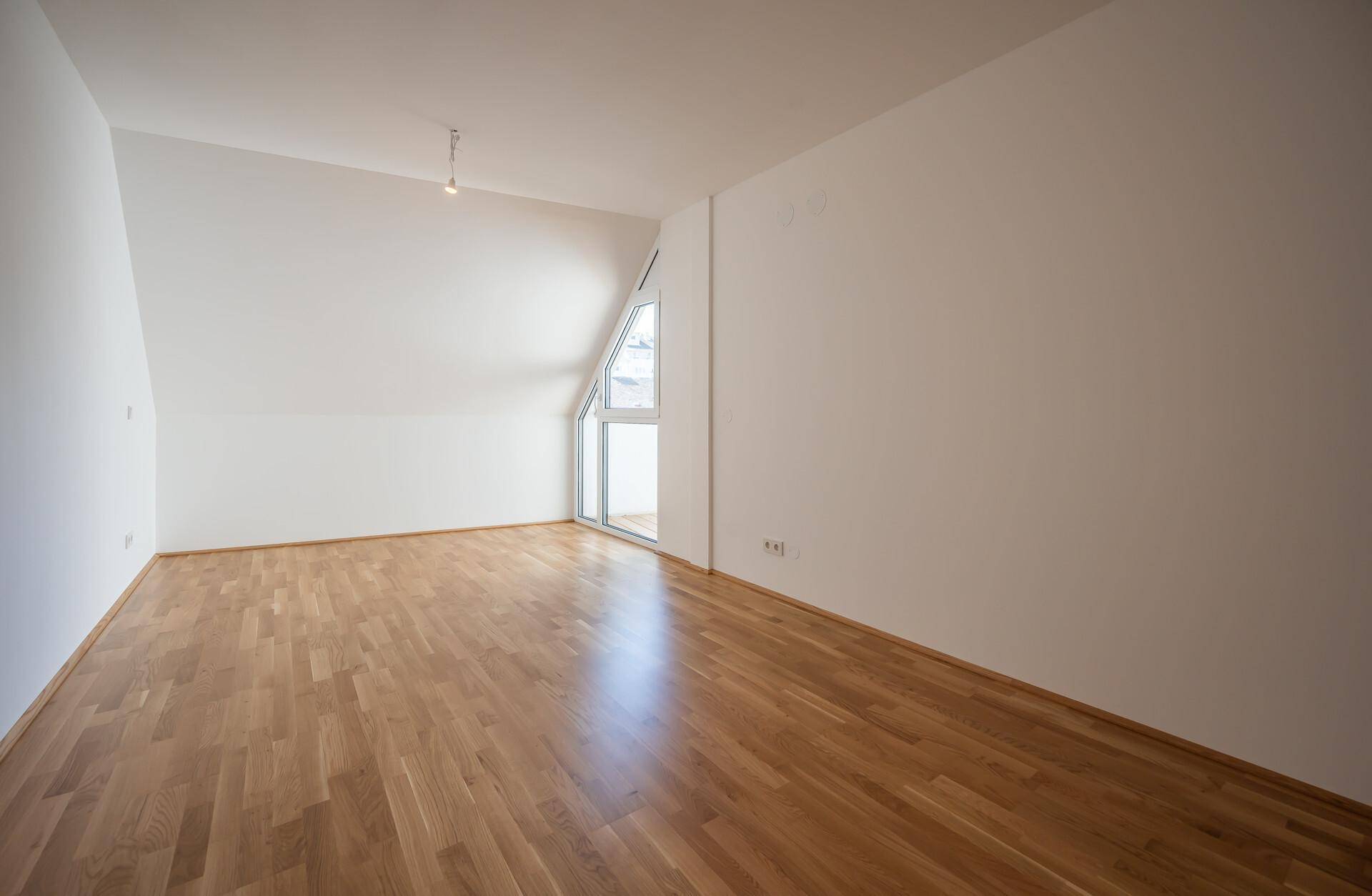 Geräumiger Raum mit Holzboden, weißen Wänden und schrägen Decken, mit Zugang zu einem Außenbereich.