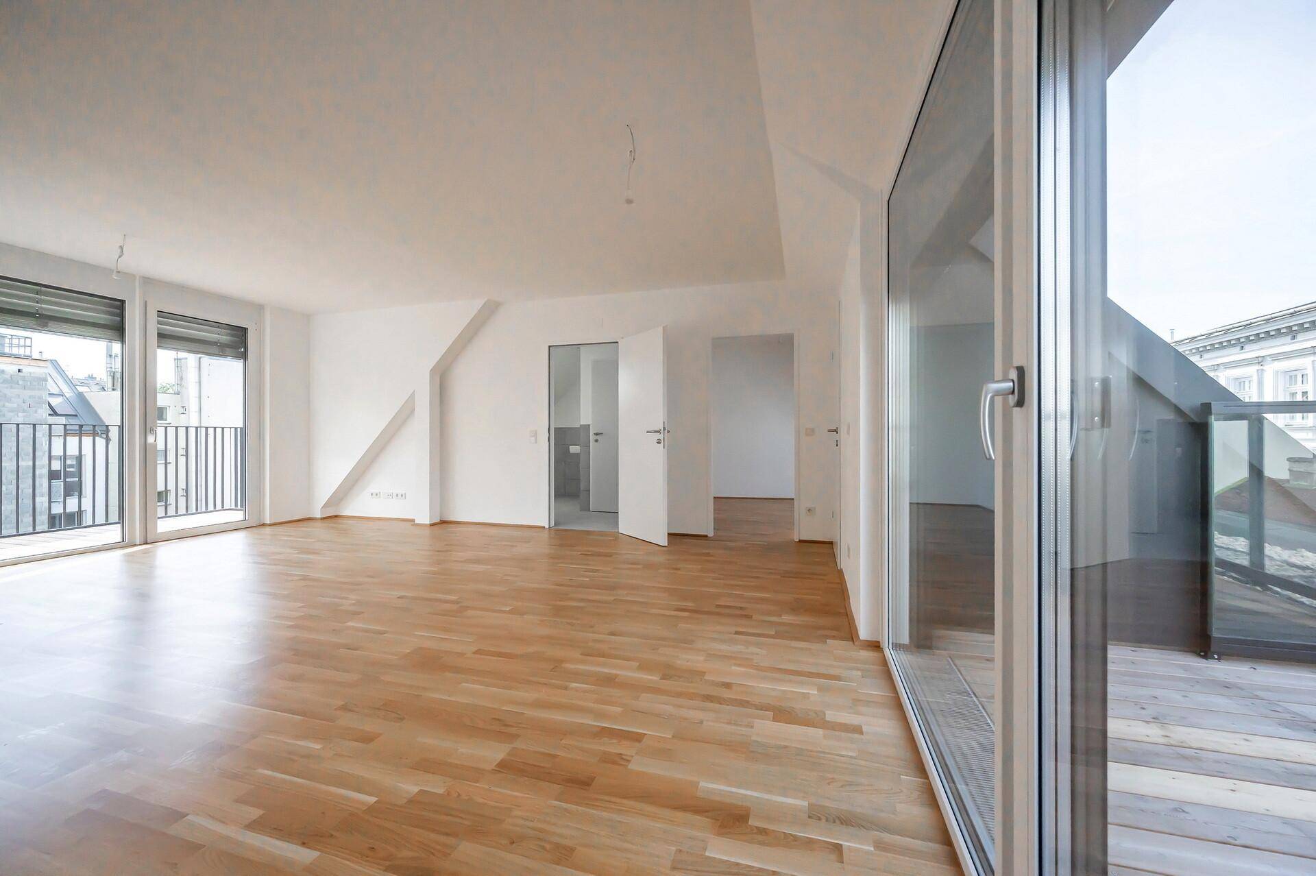 Heller, leerer Wohnraum mit Holzparkett, großen Glasfronten und Zugang zu einem kleinen Balkon.