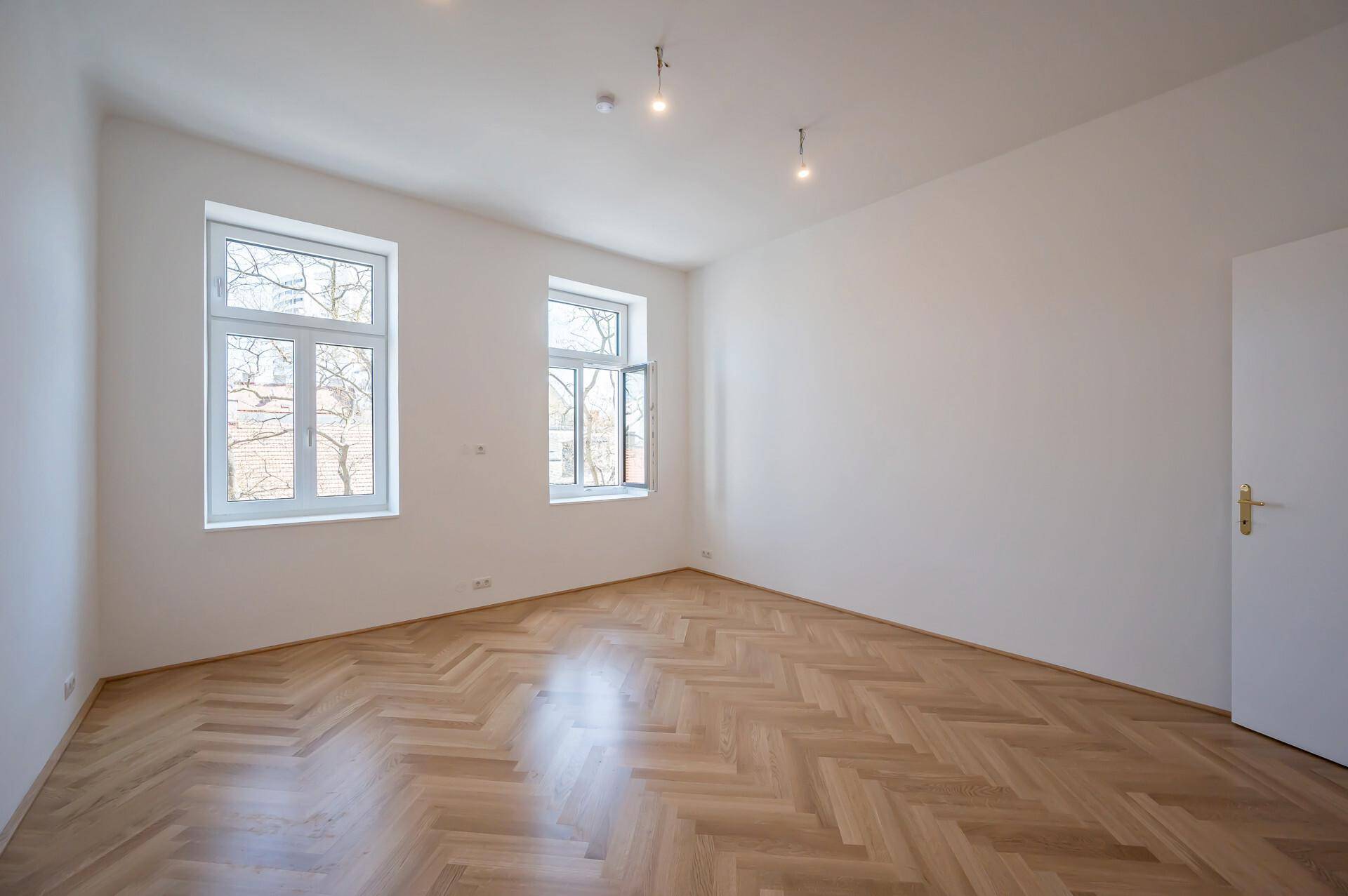 Geräumiges, leeres Zimmer mit hellem Fischgrätparkett, weißen Wänden und zwei großen Fenstern.