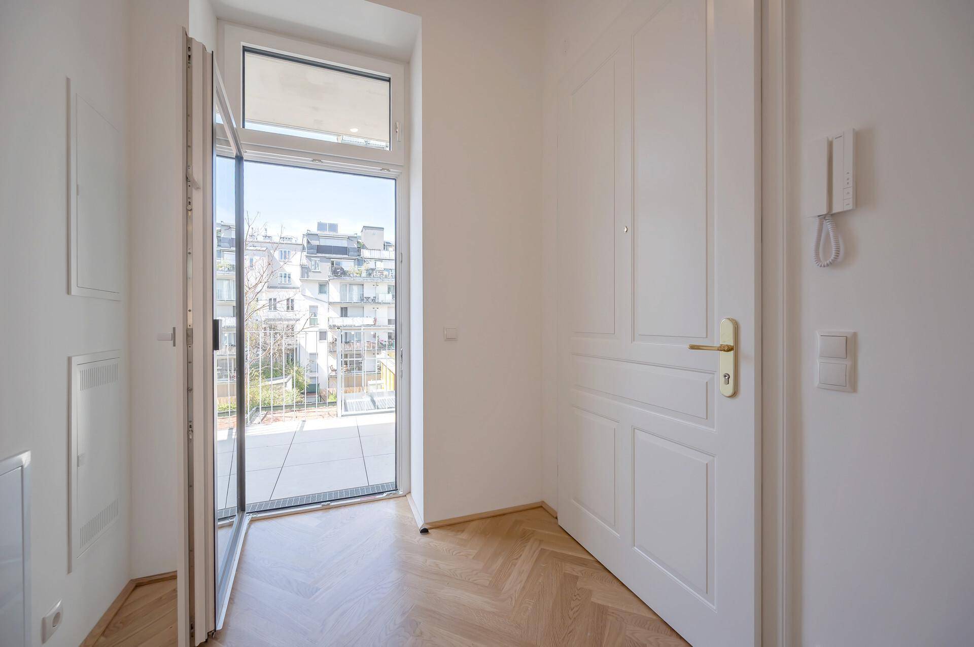 Eingangsbereich mit weißer Tür, Fischgrätparkett und Zugang zu einem Balkon oder einer Terrasse.