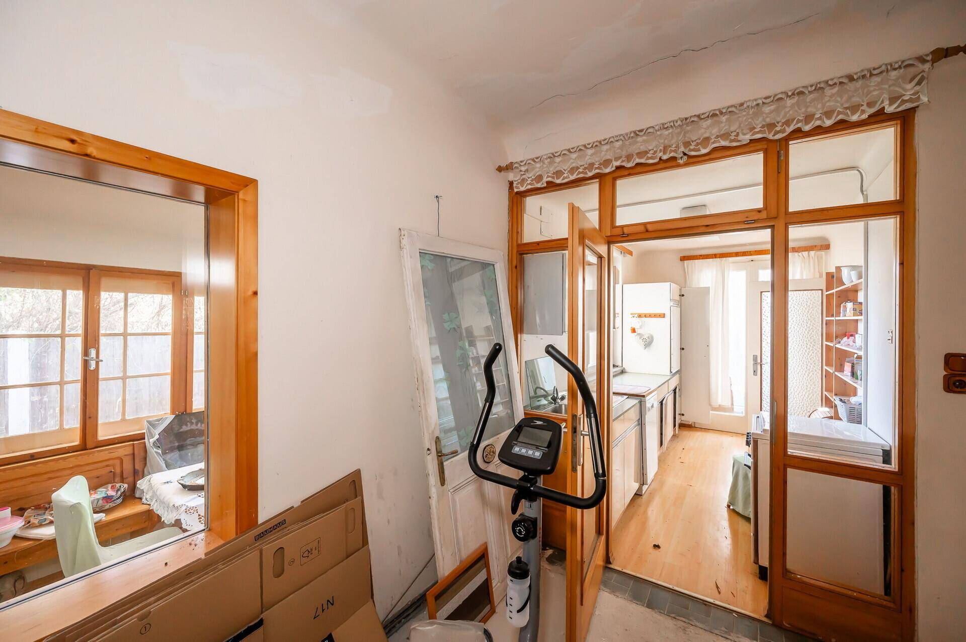 Zimmer mit Blick in die Küche und den Wintergarten, mit Fitnessgerät und Renovierungsbedarf.