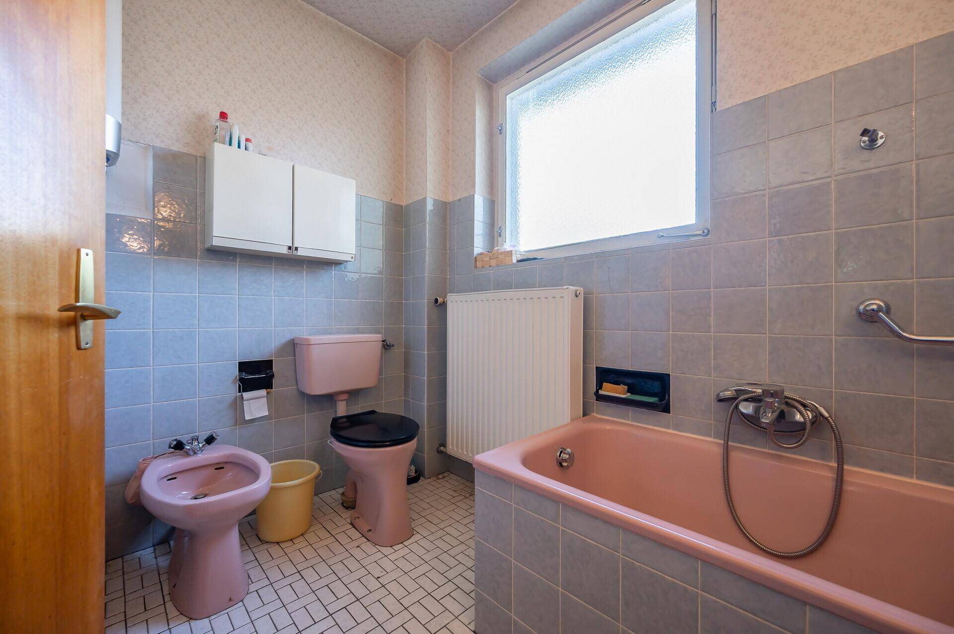 Badezimmer mit rosa Toilette, Bidet, Badewanne und grauen Wand- und Bodenfliesen.