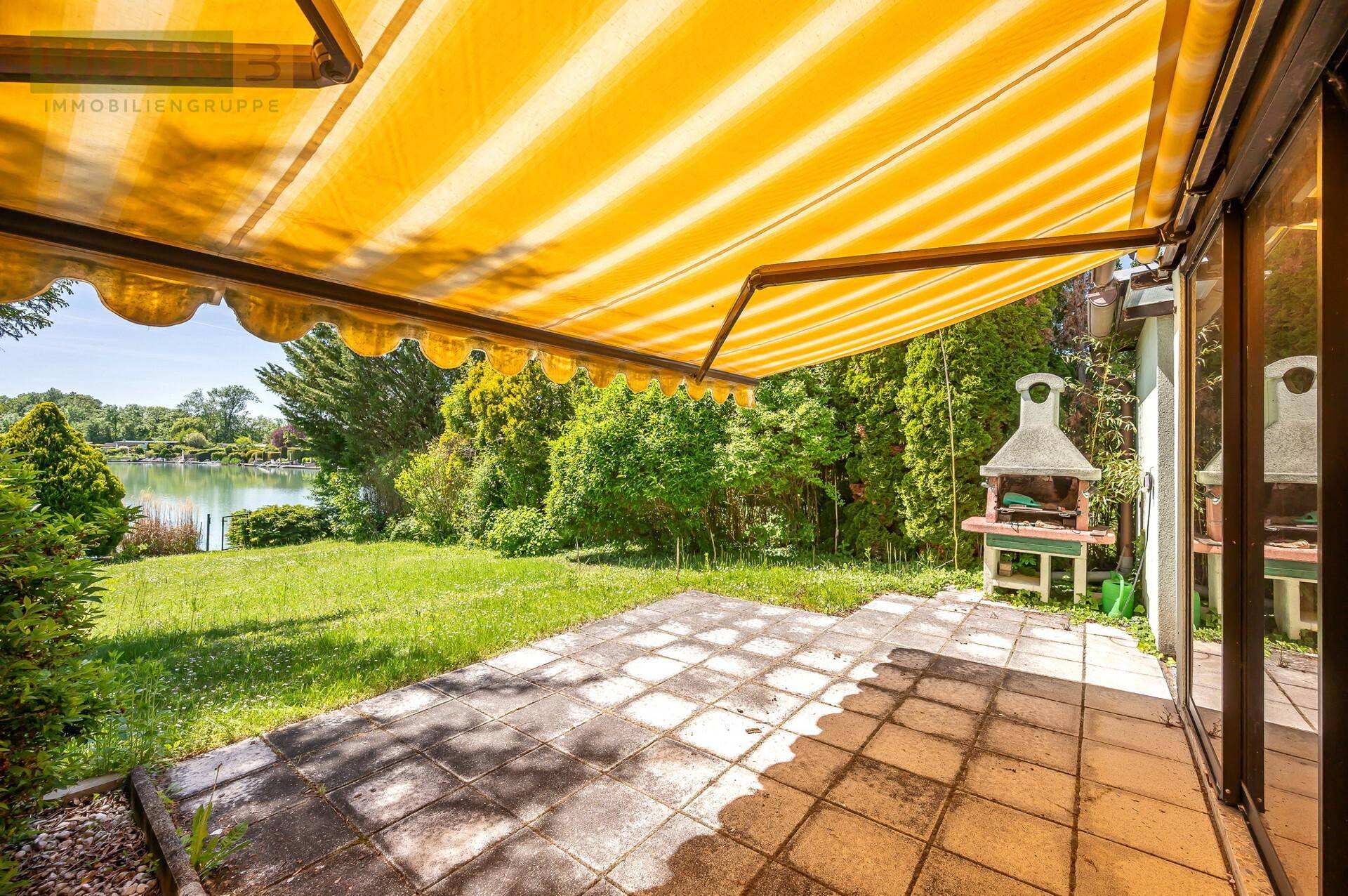 Sonnenterrasse mit gelber Markise, Grillplatz und direktem Blick auf den See und Garten.