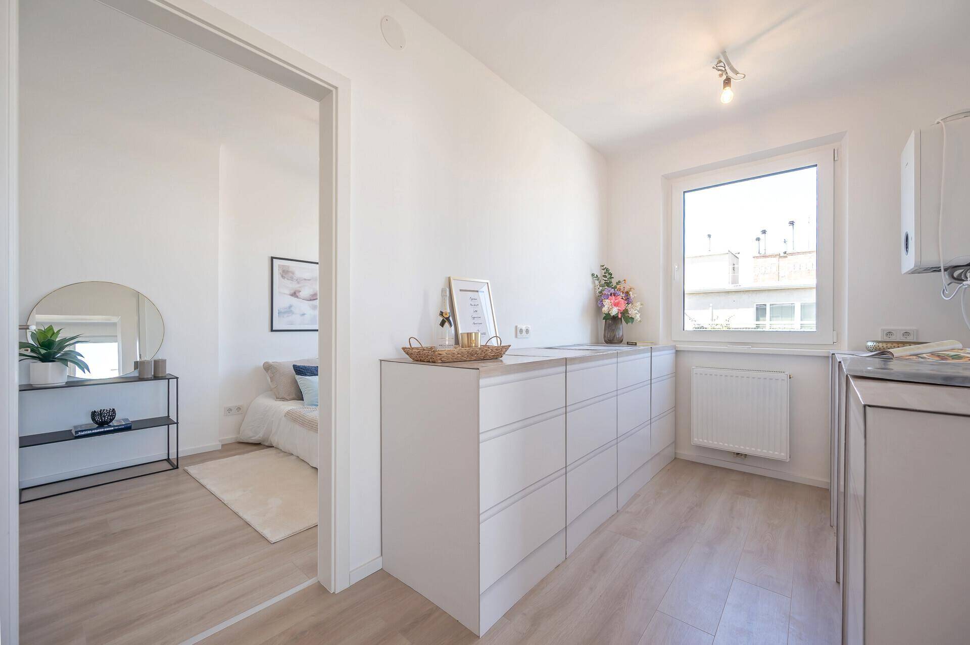 Heller Küchenbereich mit Fenster und vorbereiteten Anschlüssen, angrenzend an Schlafzimmer.