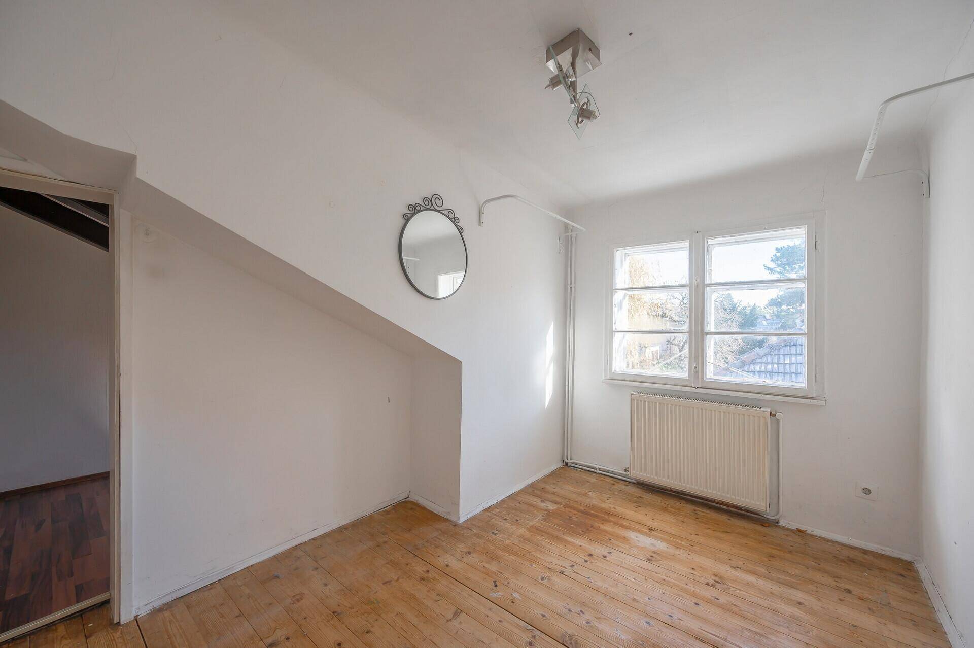 Helles Zimmer mit Holzdielenboden und Fenster, das auf die Umgebung blickt, renovierungsbedürftig.