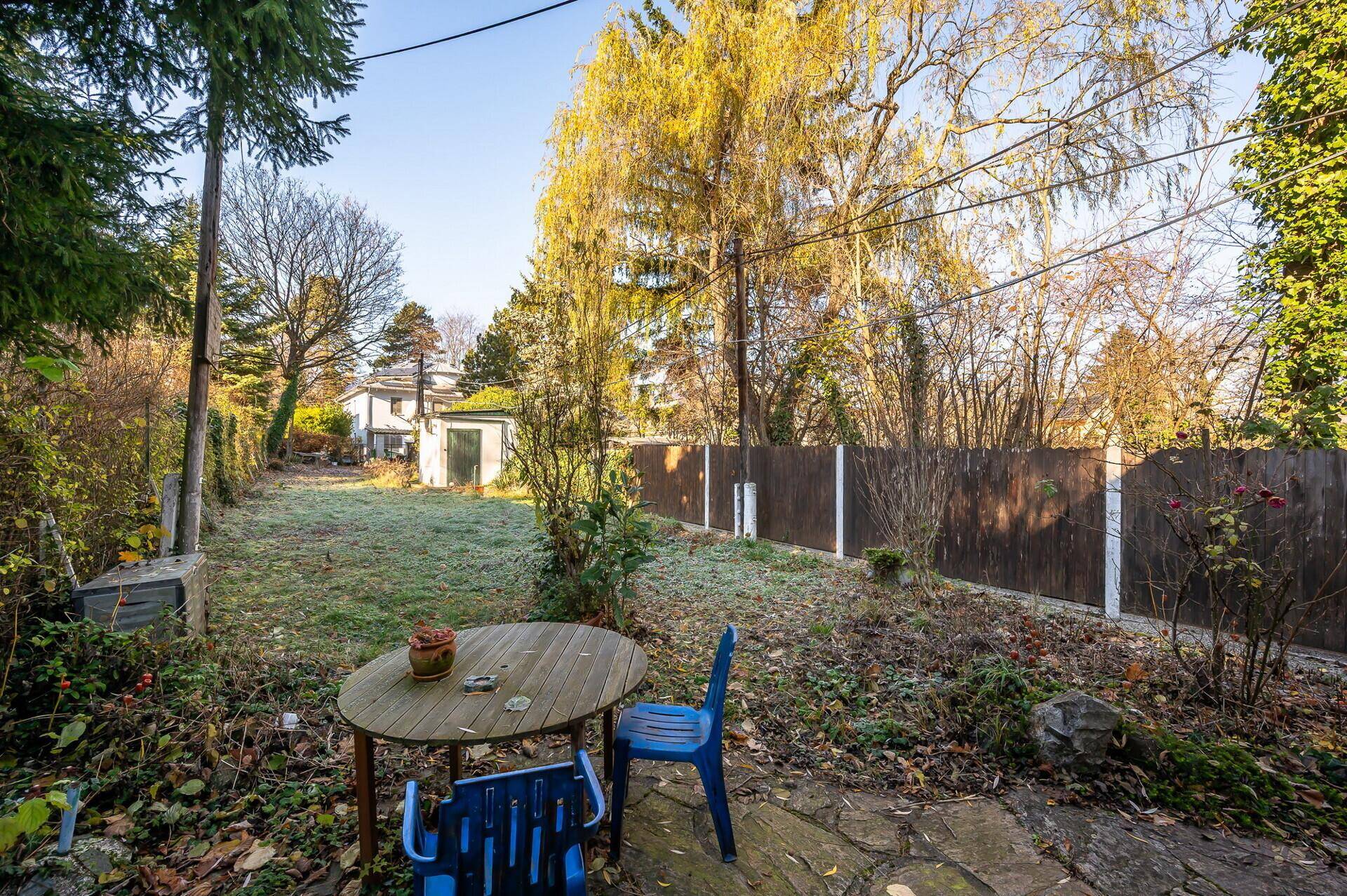 Gemütlicher Sitzbereich im Garten mit Holztisch und Stühlen, umgeben von Bäumen und Rasen.