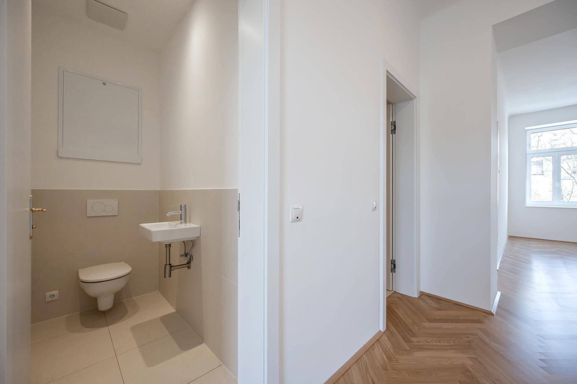 Kompaktes Gäste-WC mit wandhängender Toilette, kleinem Waschbecken und beigefarbenen Bodenfliesen.