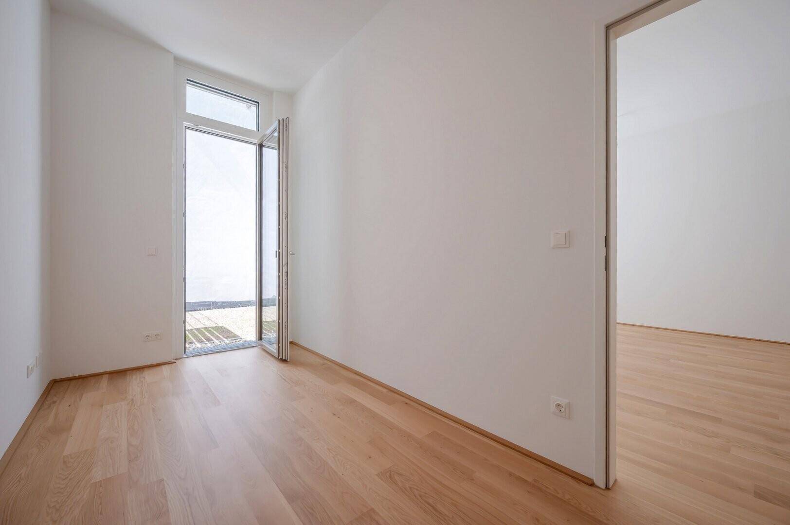 Leerer Raum mit Holzboden und großen Fenstern, die Zugang zu einem Außenbereich bieten.