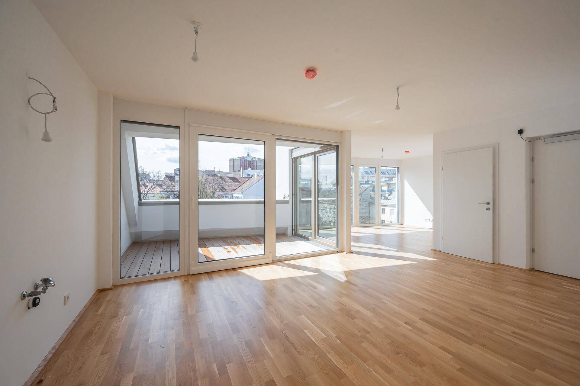 Heller Wohnraum mit Parkettboden und großen Fenstern, die viel Tageslicht hereinlassen und Zugang zum Außenbereich bieten.