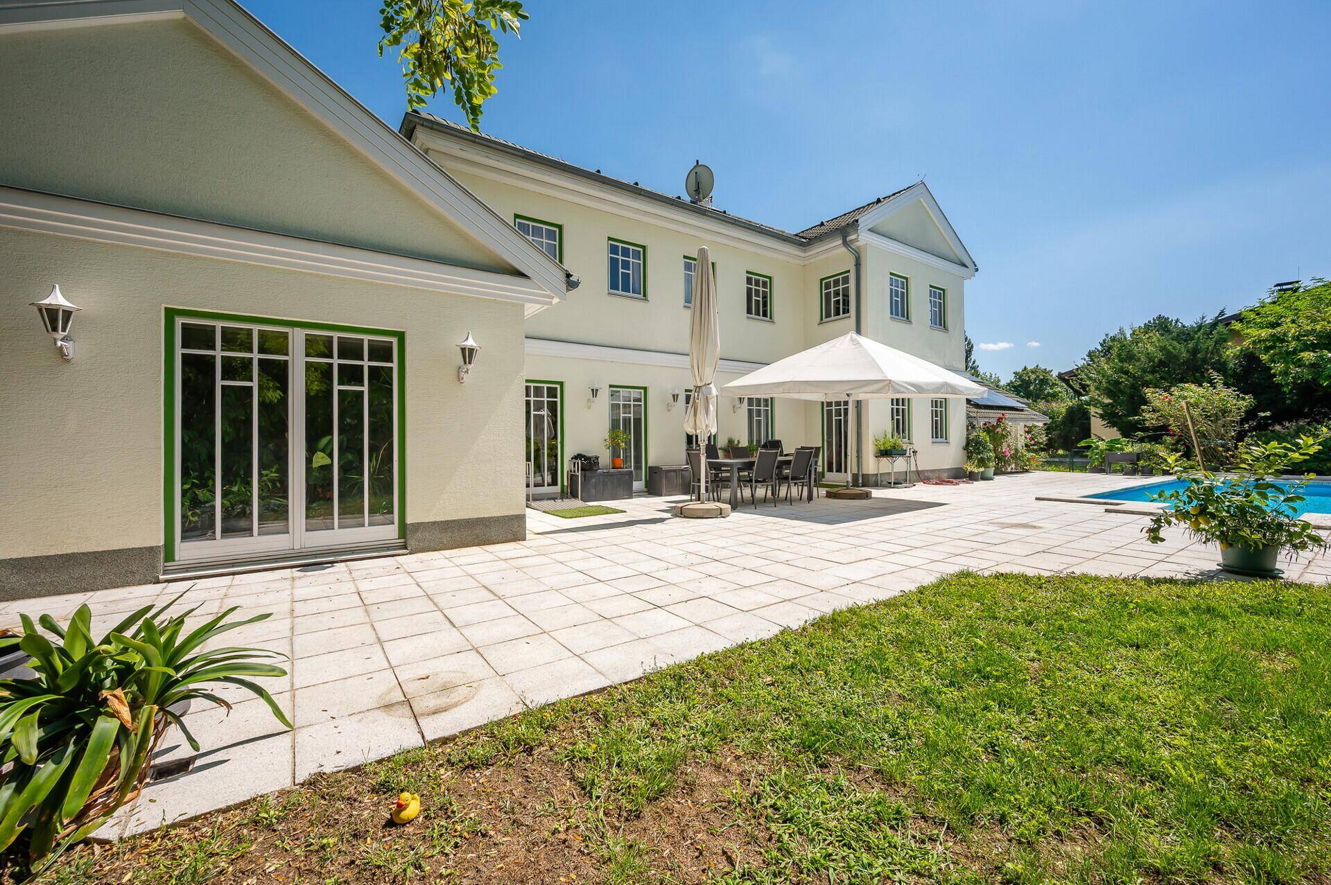 Gepflegter Gartenbereich mit Rasenfläche, Terrasse und Pool im Hintergrund.