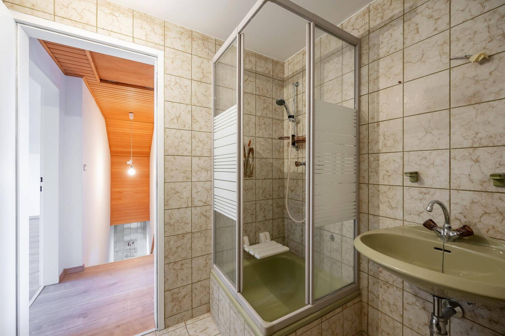 Badezimmer mit Duschkabine, Waschbecken und beigefarbenen Fliesen, verbunden mit dem Flur.