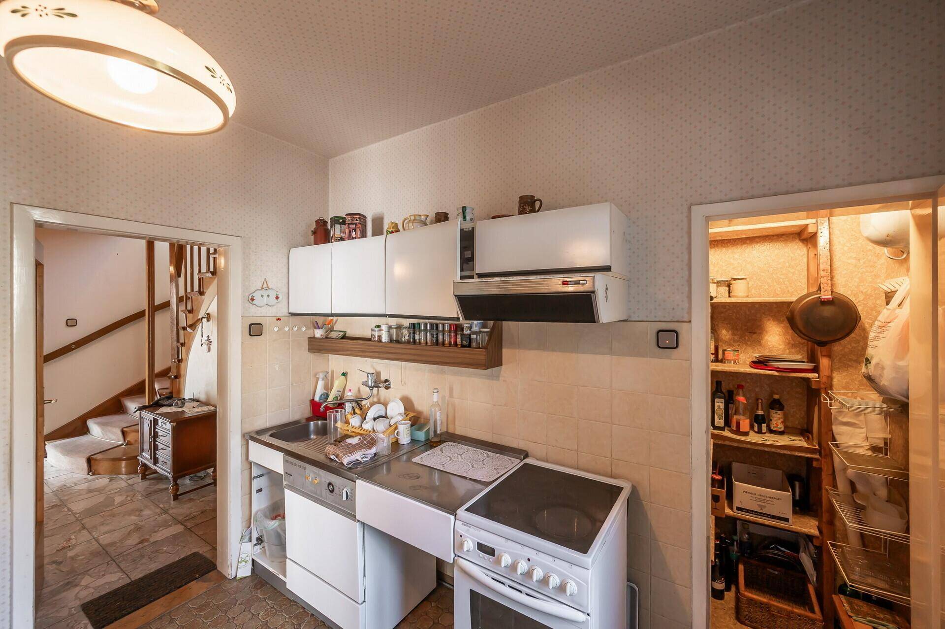 Funktionale Küche mit älteren weißen Schränken, Spüle und Herd, angrenzend an eine Speisekammer.