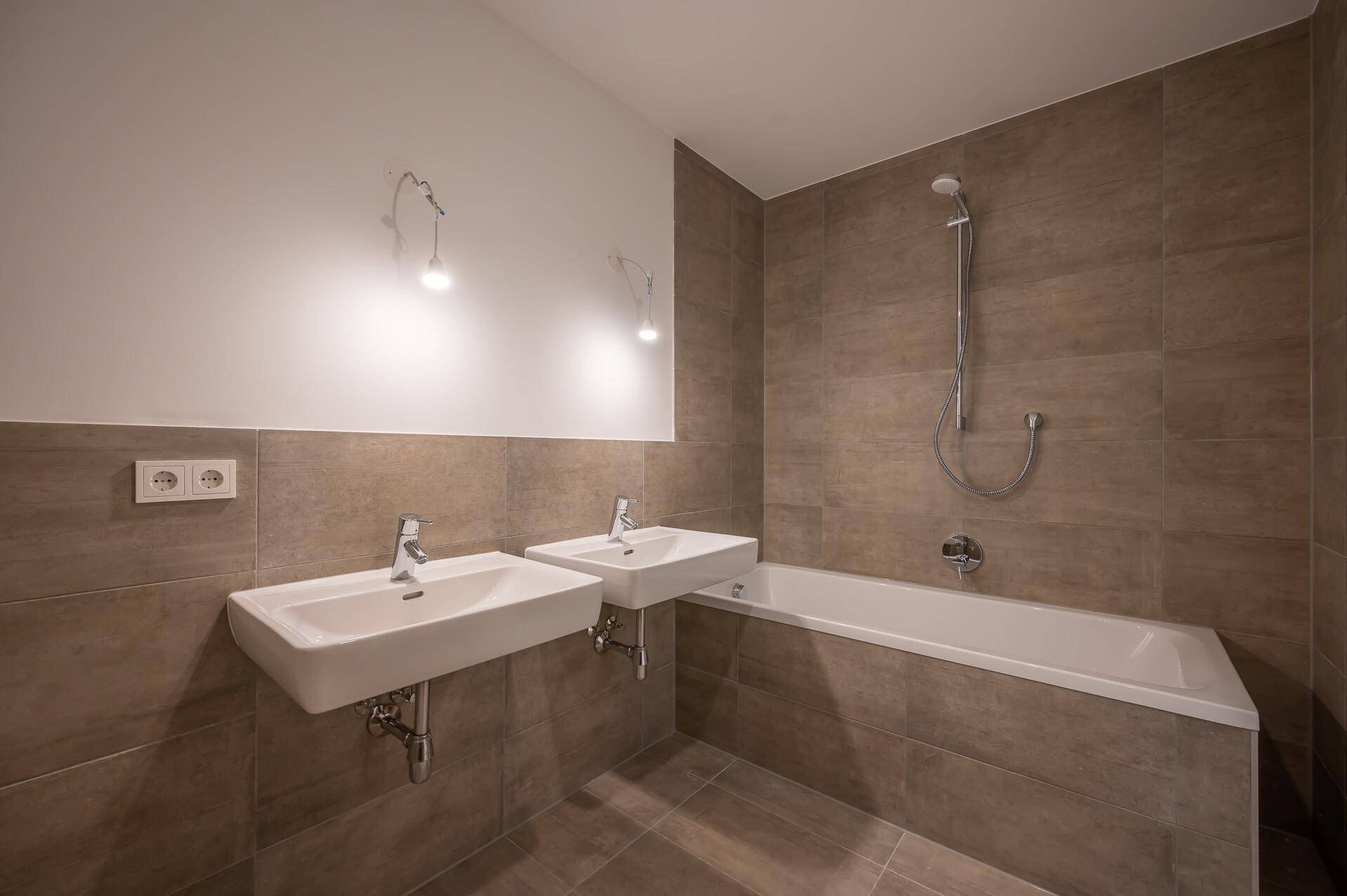 Modernes Badezimmer mit braunen Fliesen, zwei Waschbecken und einer Badewanne mit Duschfunktion.