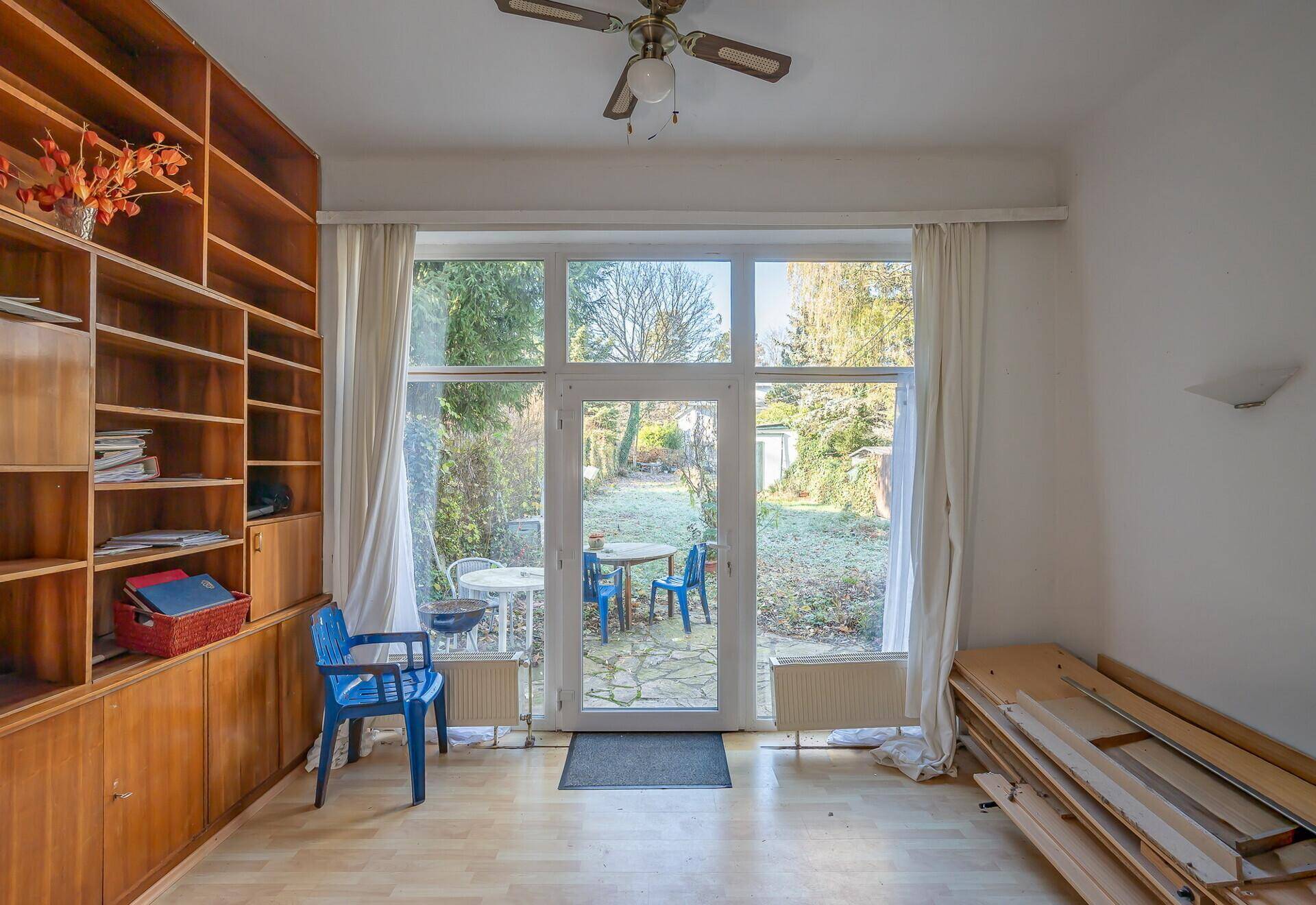 Helles Wohnzimmer mit großem Fenster zum Garten, Holzregal und Parkettboden, renovierungsbedürftig.